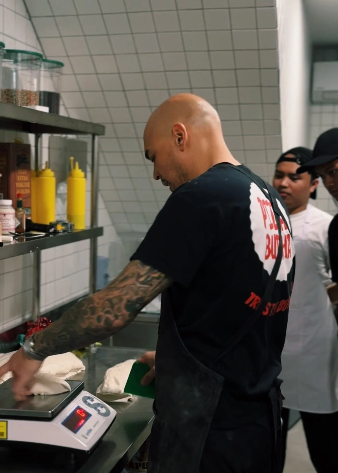 Pizza consultant Alastair Hannmann Al the Pizza Buddha training the pizza chefs. He weighs ingredients on a digital scale in a kitchen, with two other people observing.
