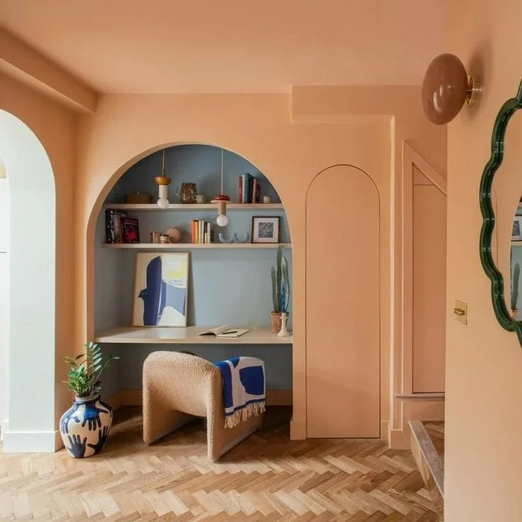 Interior view of a cozy corner with a built-in desk under an arched alcove, decorated with books, plants, and decorative items, with a plush chair and area rug, in a pastel-colored room.