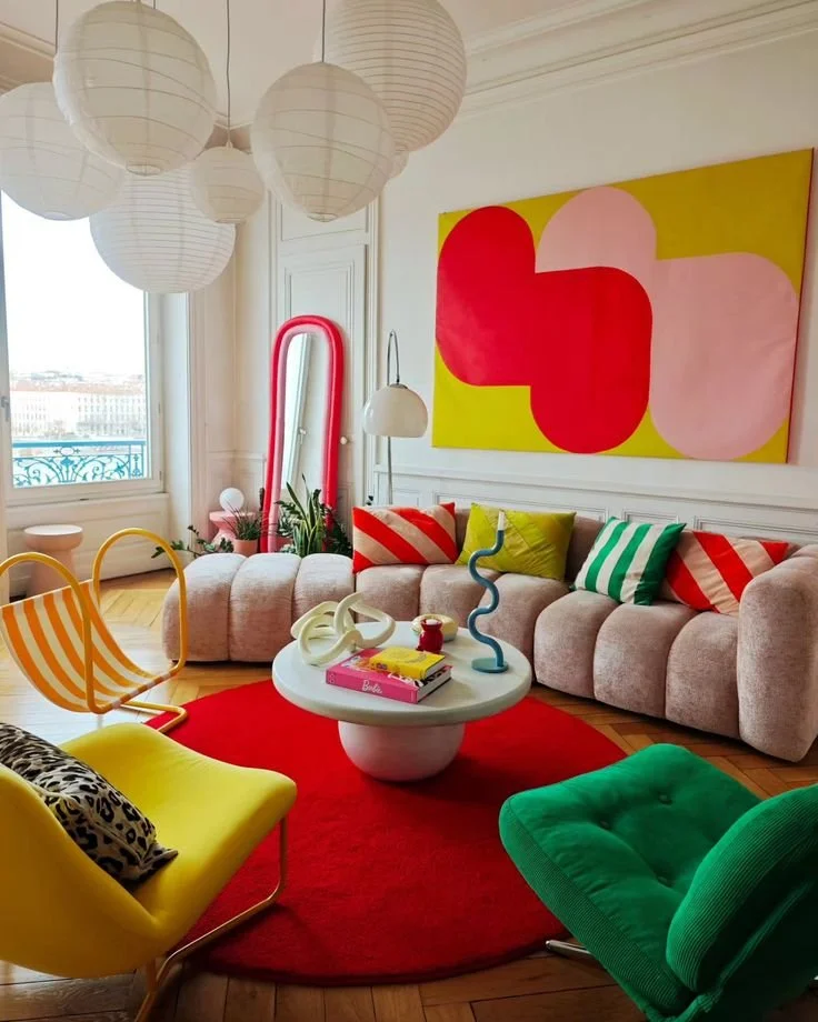 Colorful modern living room with pastel pink sofa, yellow and green chairs, abstract artwork, white pendant lights, and a red area rug.
