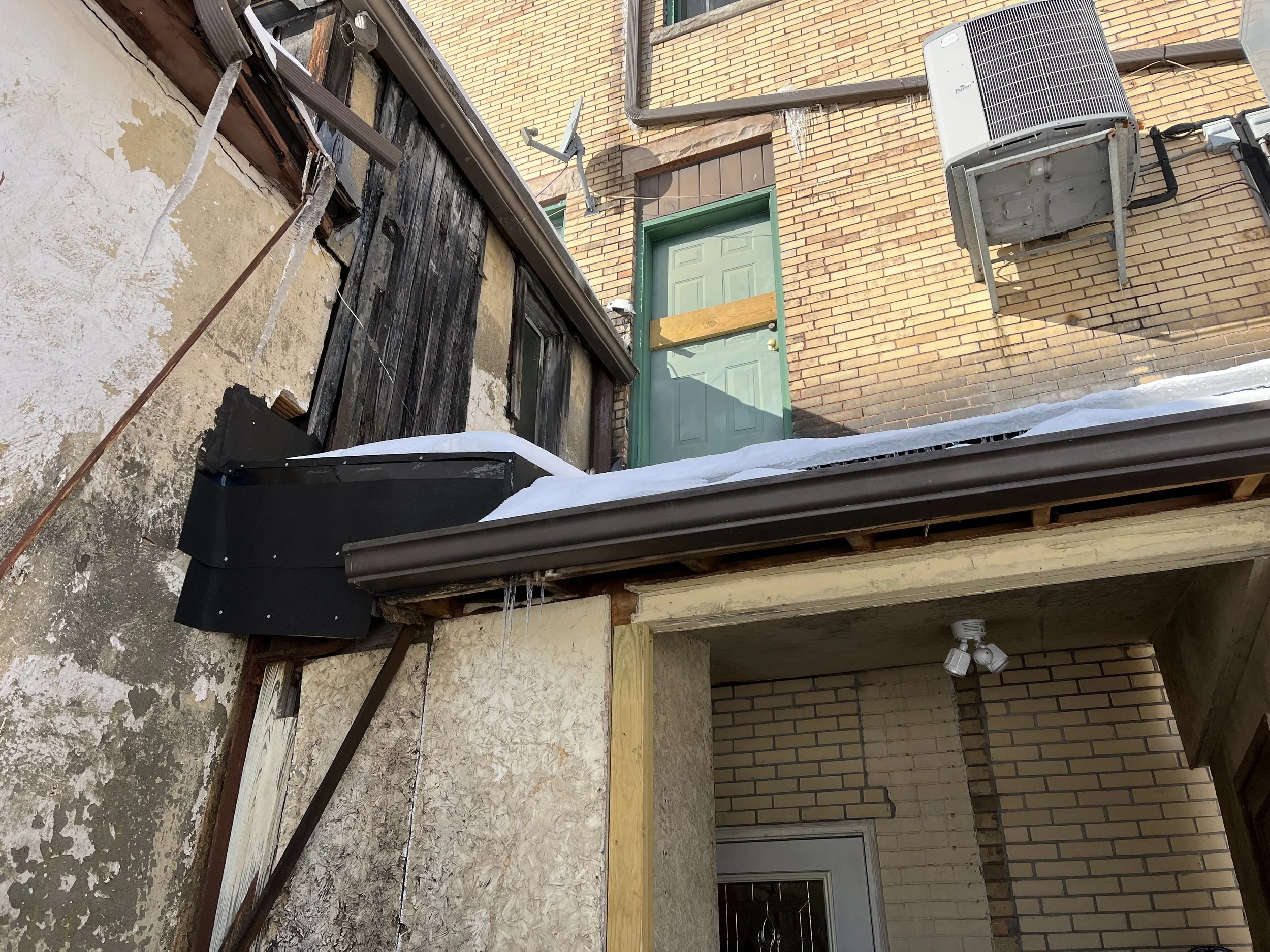 Exterior view of a building with damaged siding, snow-covered roof, and icicles.