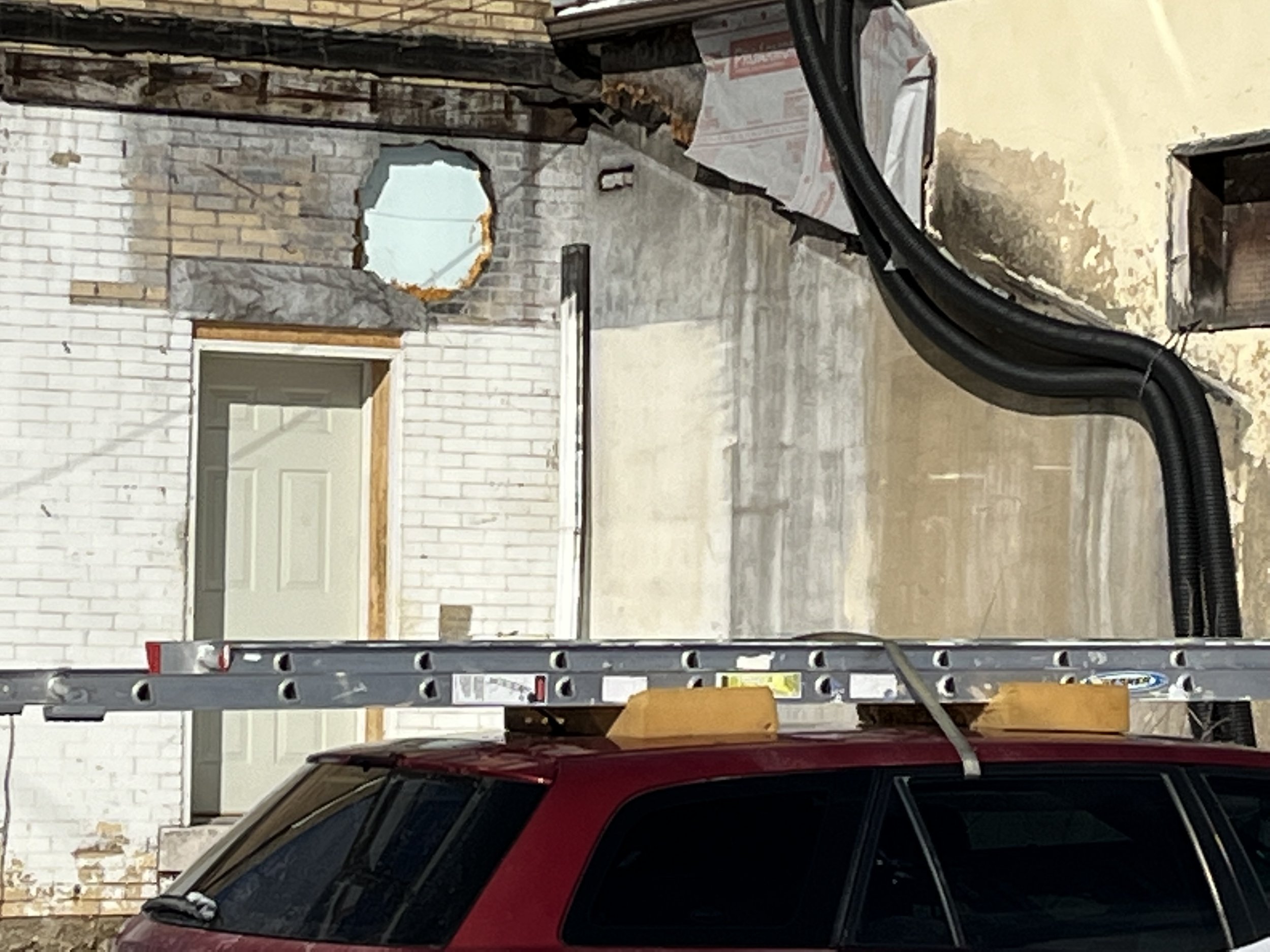 Exterior wall with a partially exposed hole and a white door. A red vehicle is parked with a ladder on top. Black pipes run along the wall.
