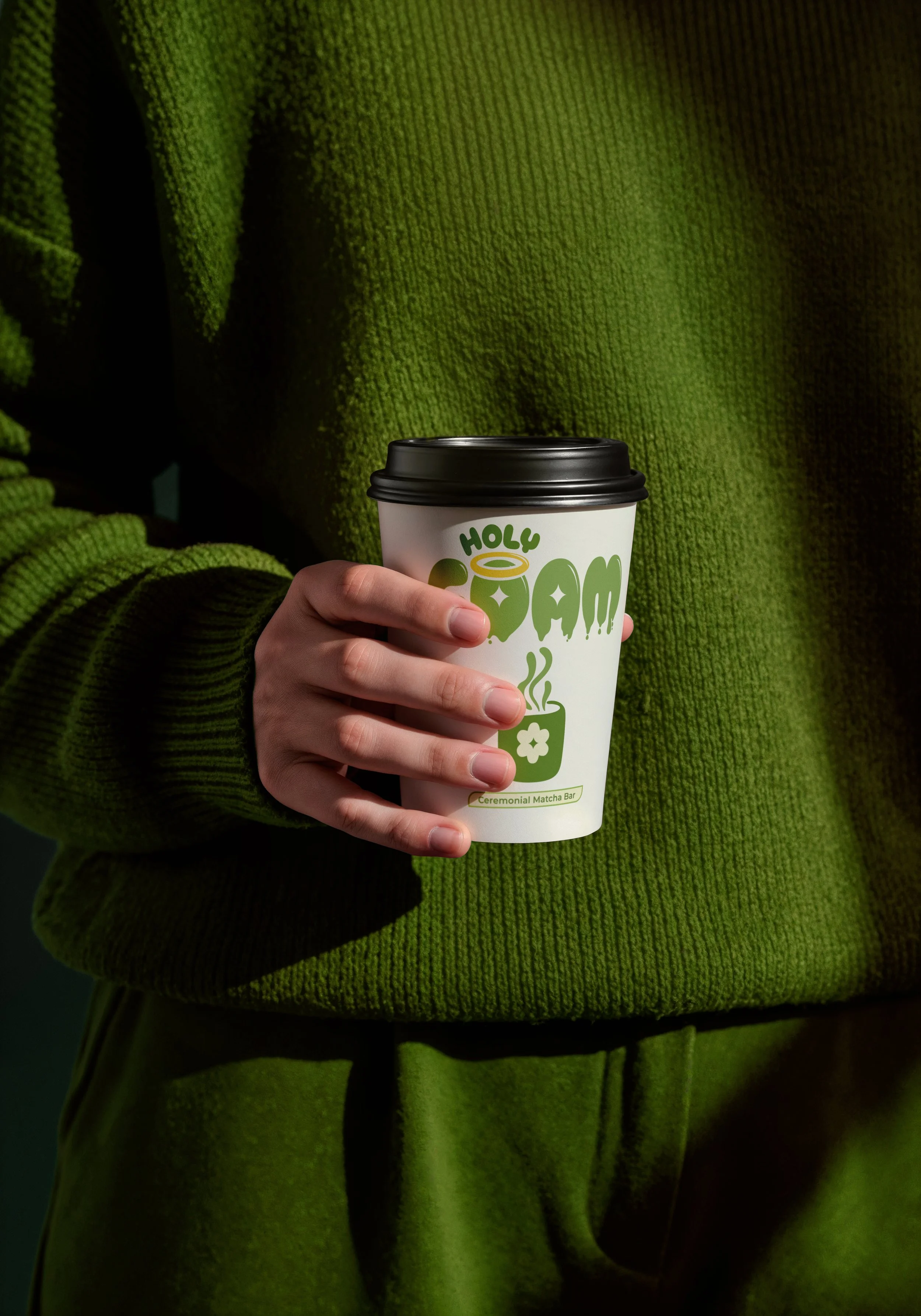 Persona con suéter verde sosteniendo un vaso desechable de té con el logo 'Holy Tea', que indica una barra de té ceremonial de matcha.