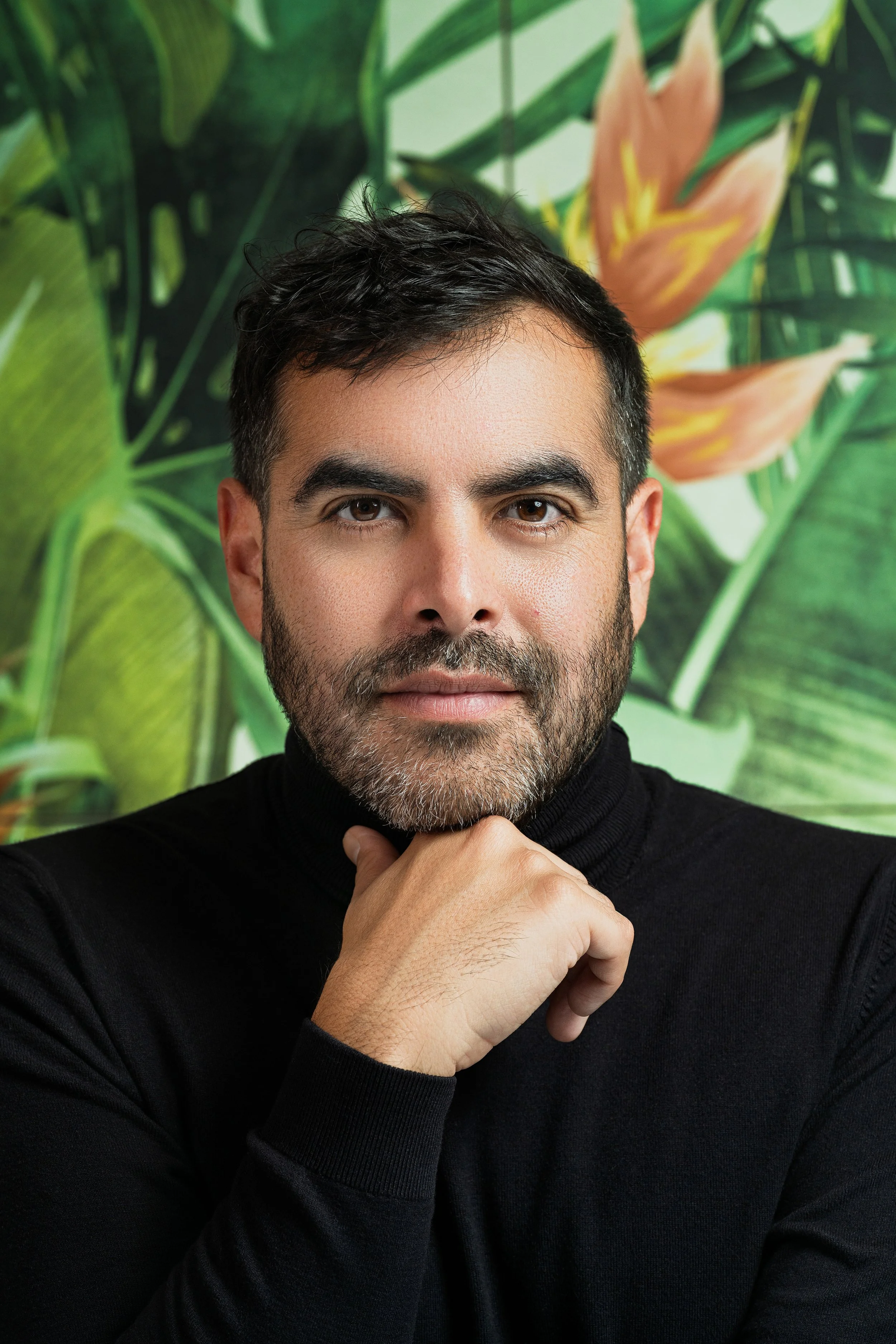 Retrato de un hombre con barba y cabello oscuro, que lleva puesta una suéter negro, con fondo de plantas con hojas verdes y una flor color durazno, en un entorno natural o decorativo.