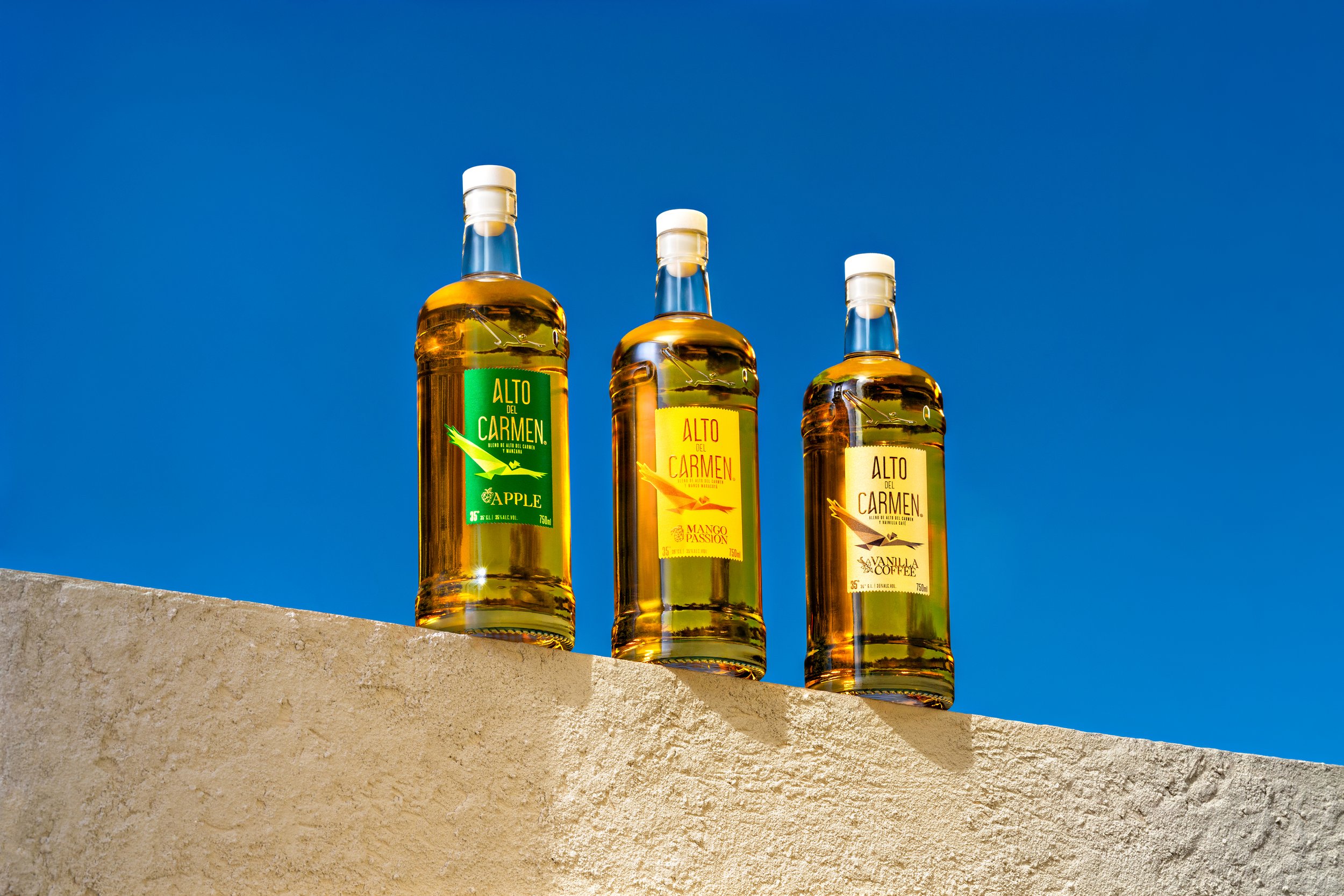Tres botellas de licor con diferentes etiquetas de sabores, sobre una superficie de piedra, con cielo azul de fondo.