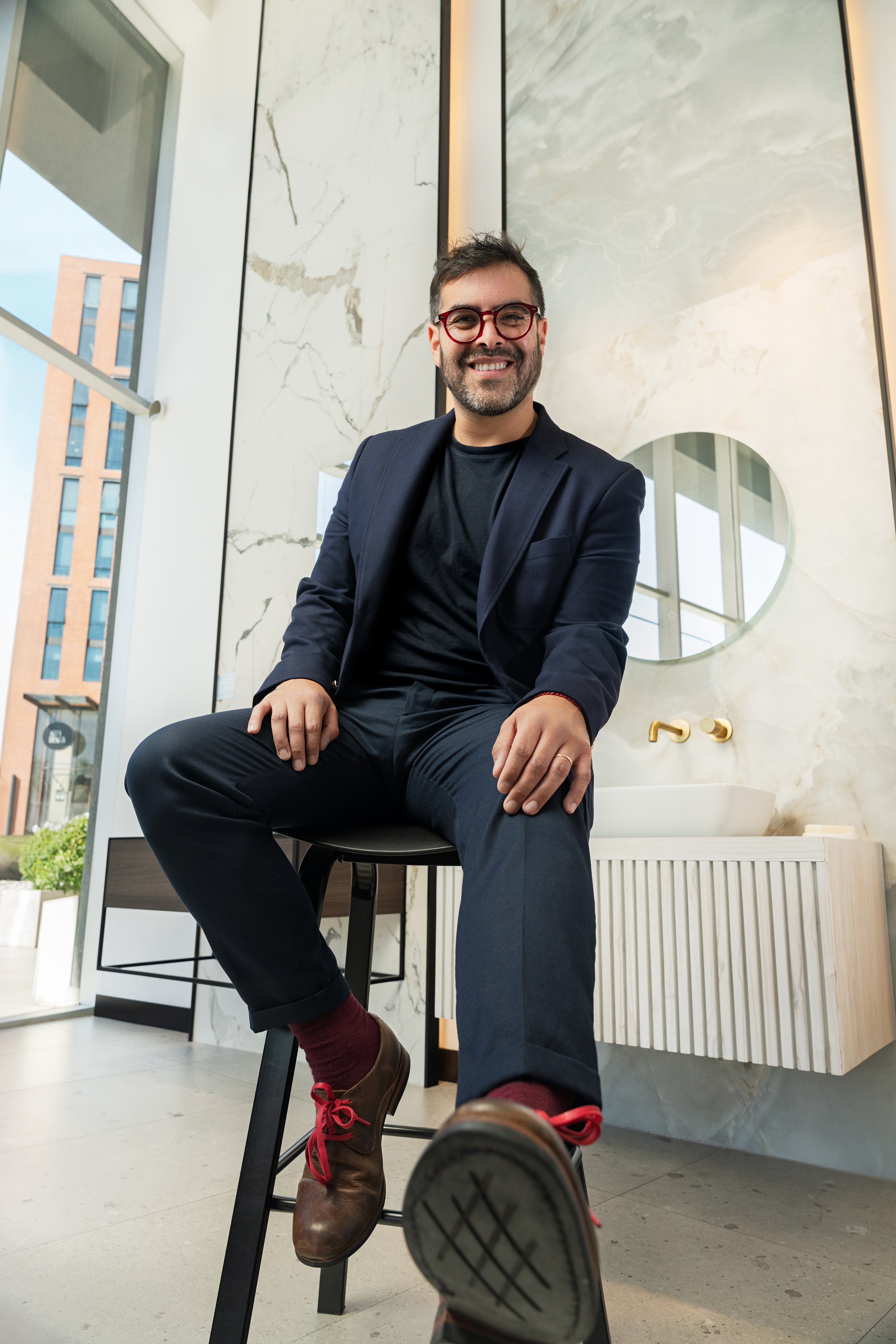 Hombre sonriente con barba, lentes rojos y chaqueta azul, sentado en un bar con fondo de paredes de mármol y ventanas grandes, en un ambiente moderno y luminoso.