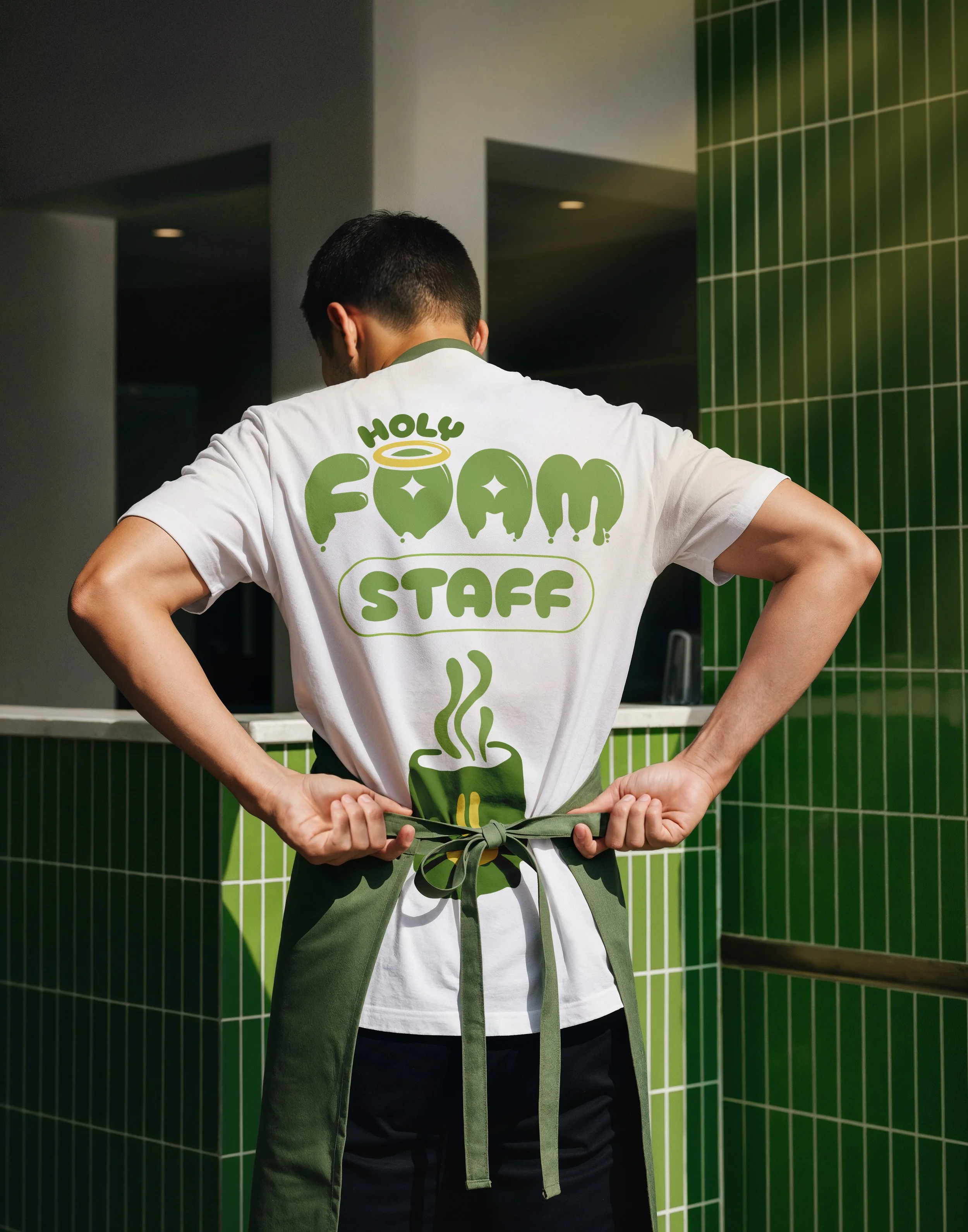 Persona vista de espaldas usando una camiseta blanca que dice 'Holy Foam Staff' con un diseño de una taza con vapor y un halo, en un espacio con paredes verdes y blancas.