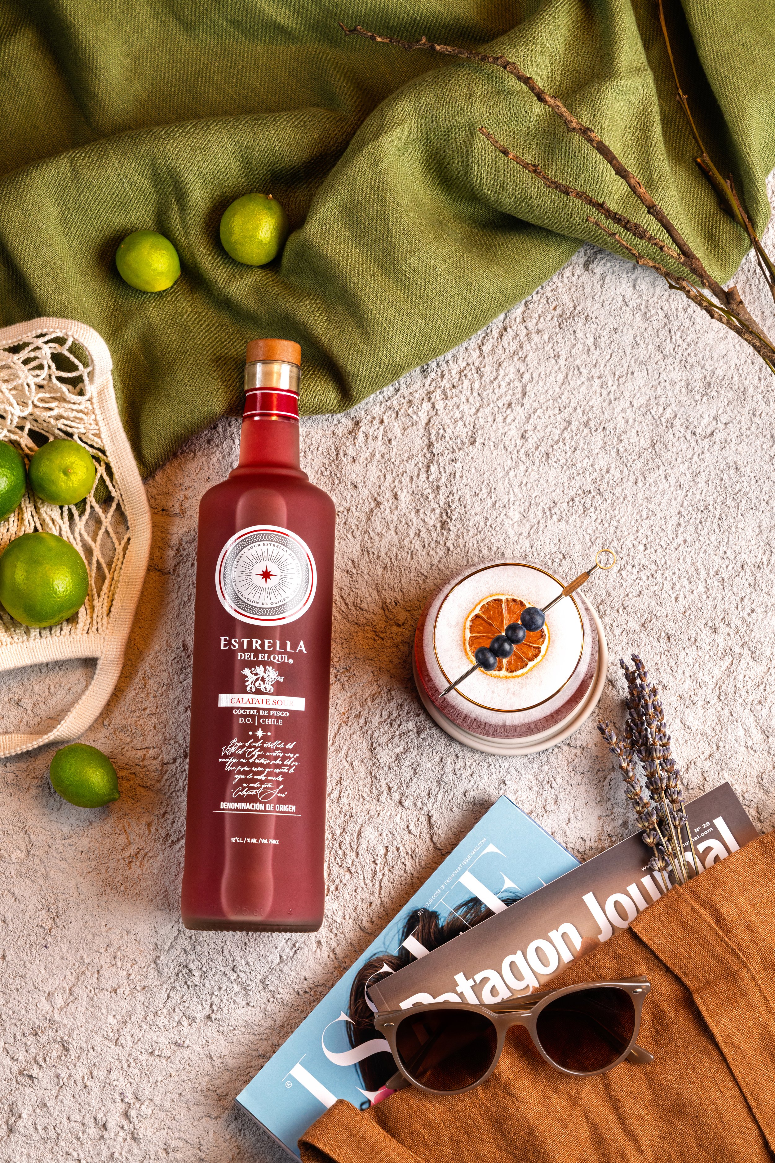 Botella de licor de color burdeos sobre una superficie, rodeada de limones verdes, un libro con gafas de sol y una revista, con flores de lavanda y ramas secas, y una tela naranja