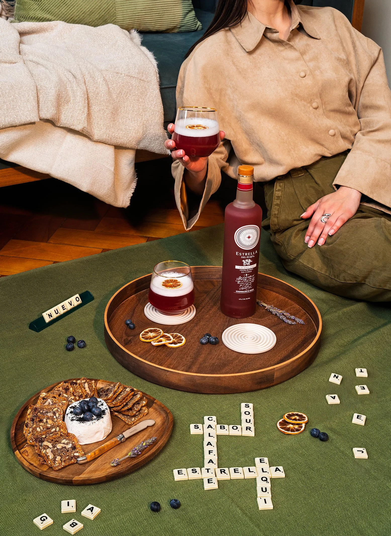 Seis vasos con bebida de color rojo, decorados con rodajas de naranja seca, en una mesa de madera. También hay diferentes tipos de galletas y postres, una botella de licor y letras de juegos de palabras en bloques blancos, con palabras en español, en