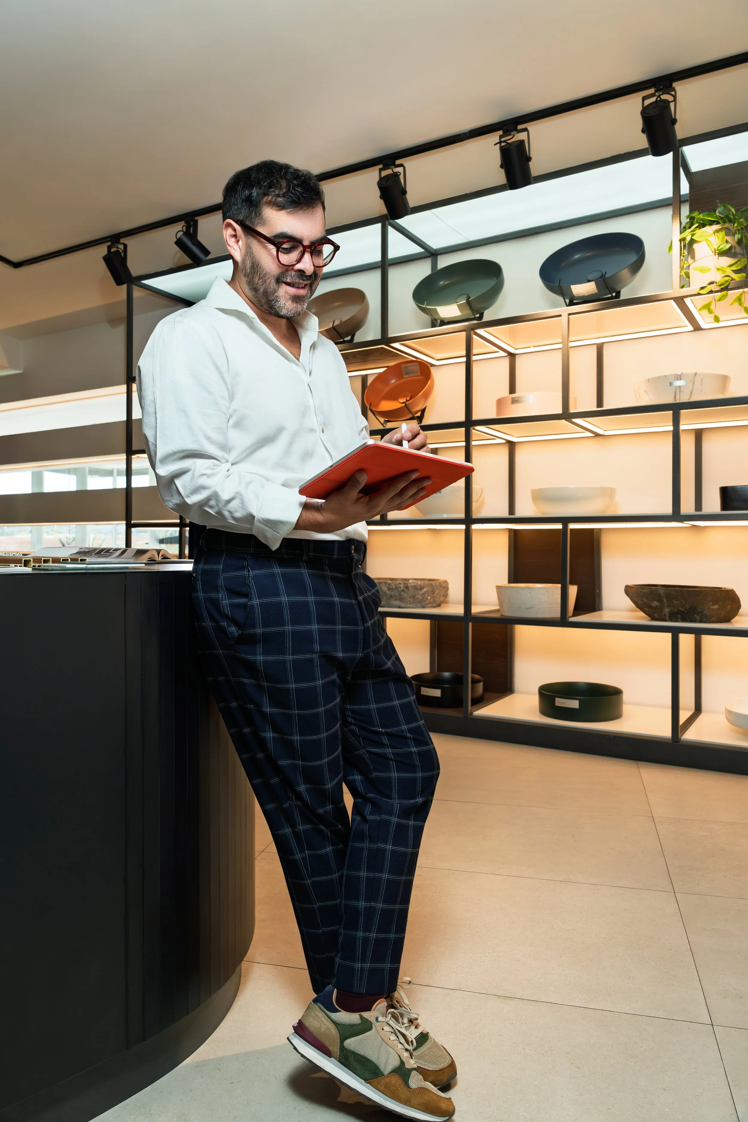 Hombre con barba y gafas de montura roja, viste camisa blanca, pantalones a cuadros y zapatos deportivos, apoyado en una mesa de exhibición, en una tienda de muebles, rodeado de estantes con platos y cuencos de cerámica y en un ambiente moderno con i