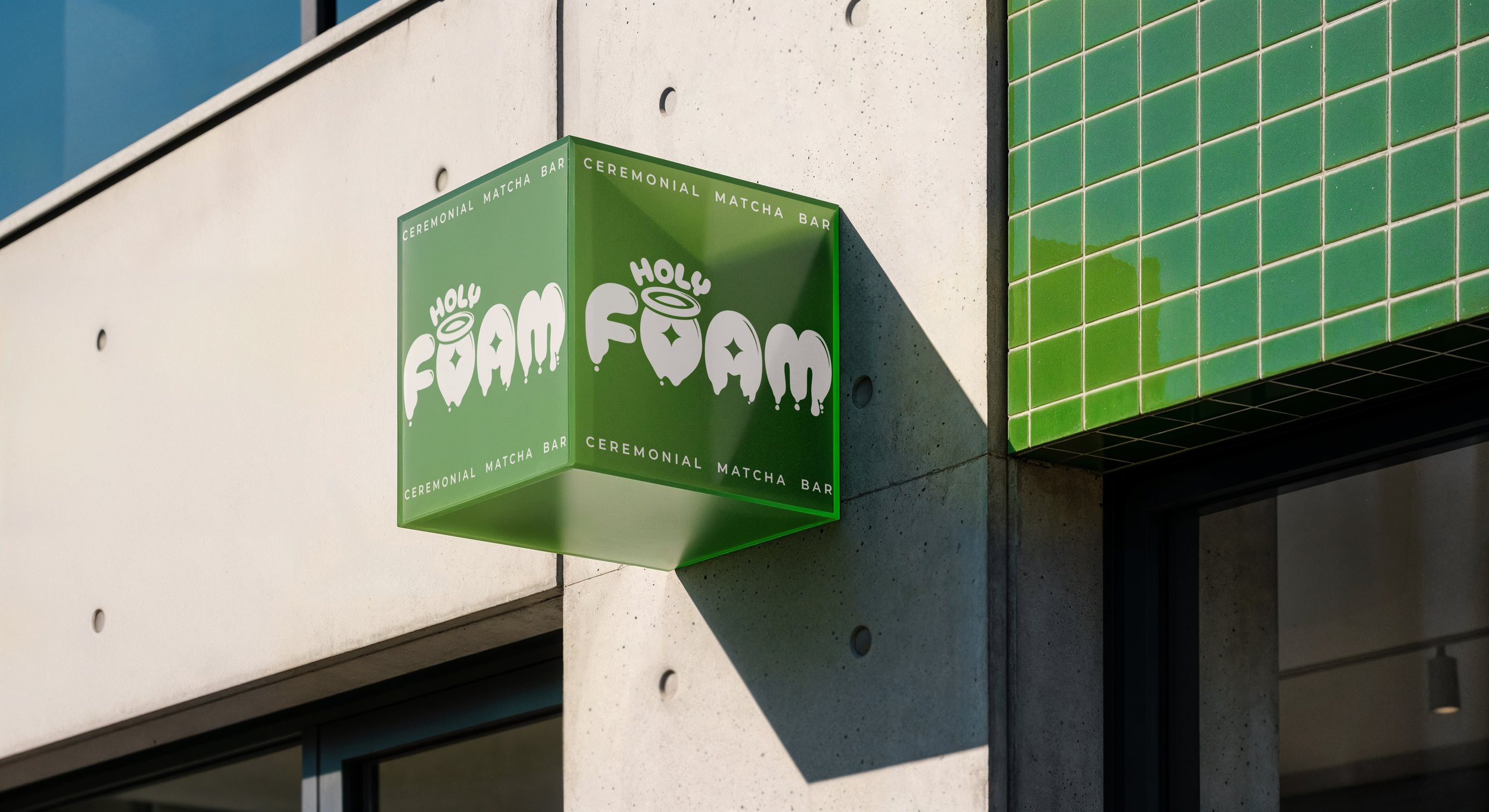 Cartel de un bar de té de matcha llamado Holy FŌAM, en color verde con letras blancas, en la fachada de un edificio moderno con paredes de concreto y detalles en verde.