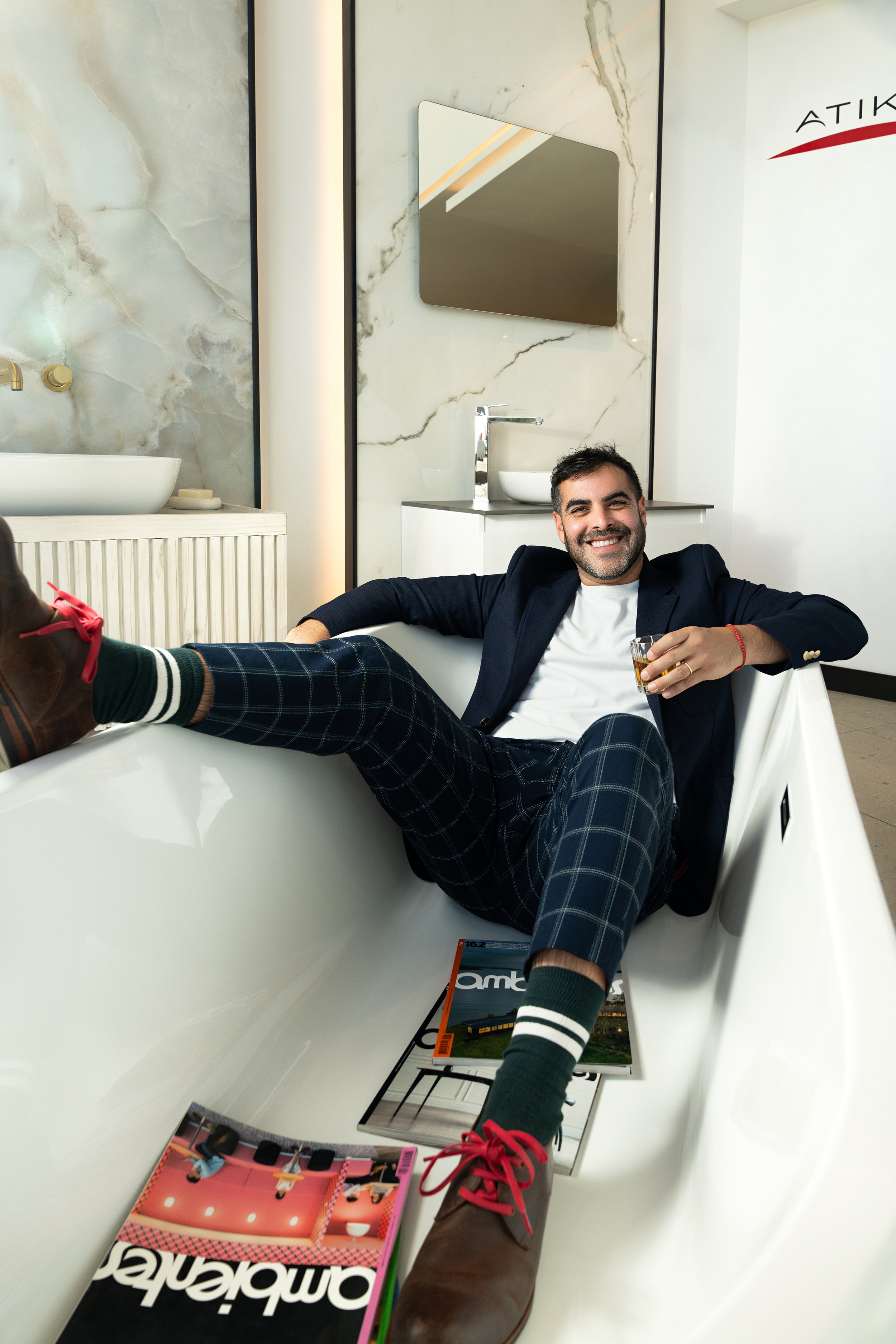 Hombre sonriendo en una bañera moderna, vestido formal con blazer, pantalones a cuadros y zapatillas rojas con calcetas con rayas, sosteniendo un vaso de bebida. Hay revistas en el suelo y un espejo y lavabo en el fondo con paredes de mármol.