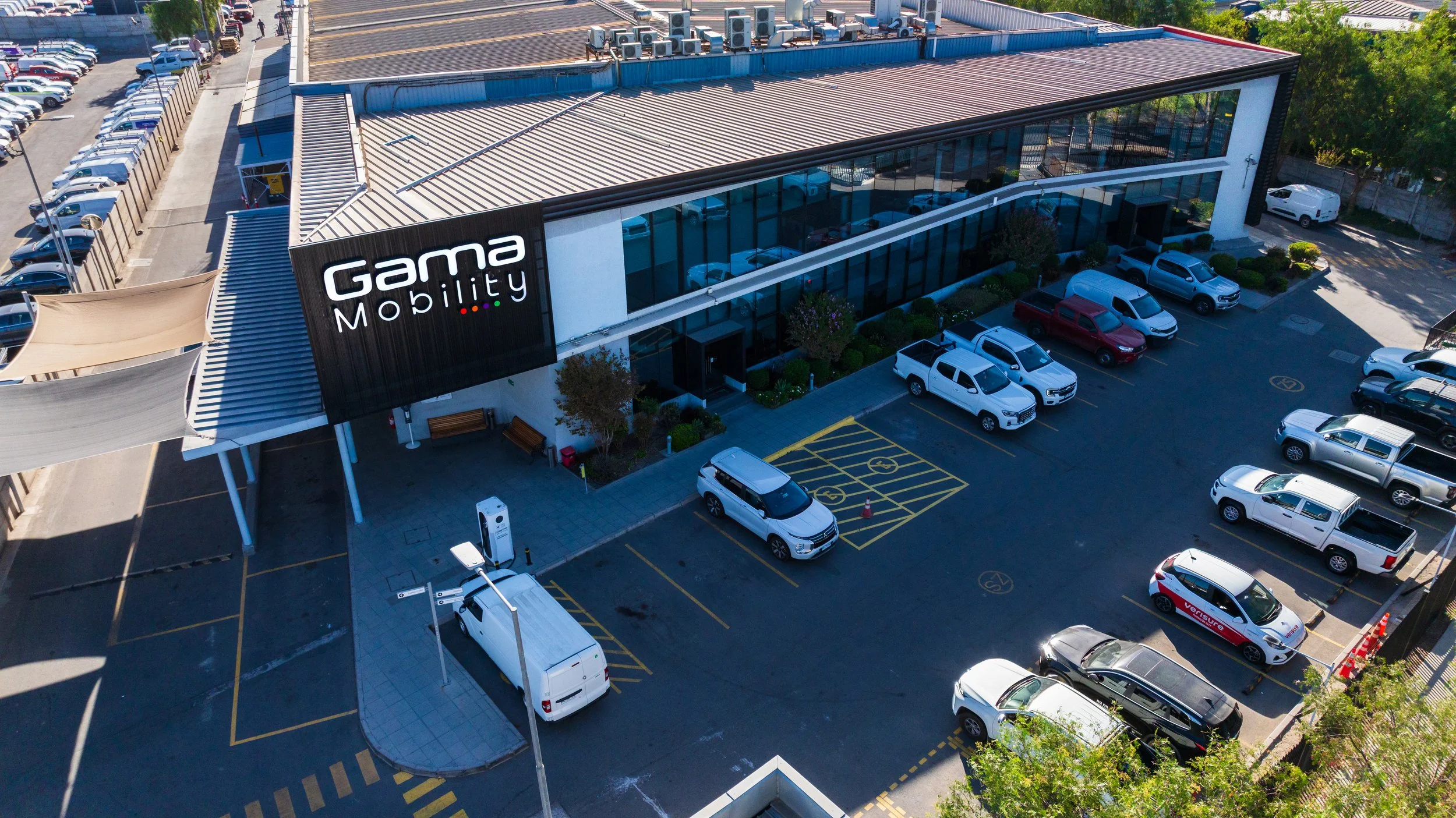 Vista aérea de un edificio con letrero que dice 'Gama Mobility', rodeado de un estacionamiento con varios autos. Hay árboles y una calle al fondo.