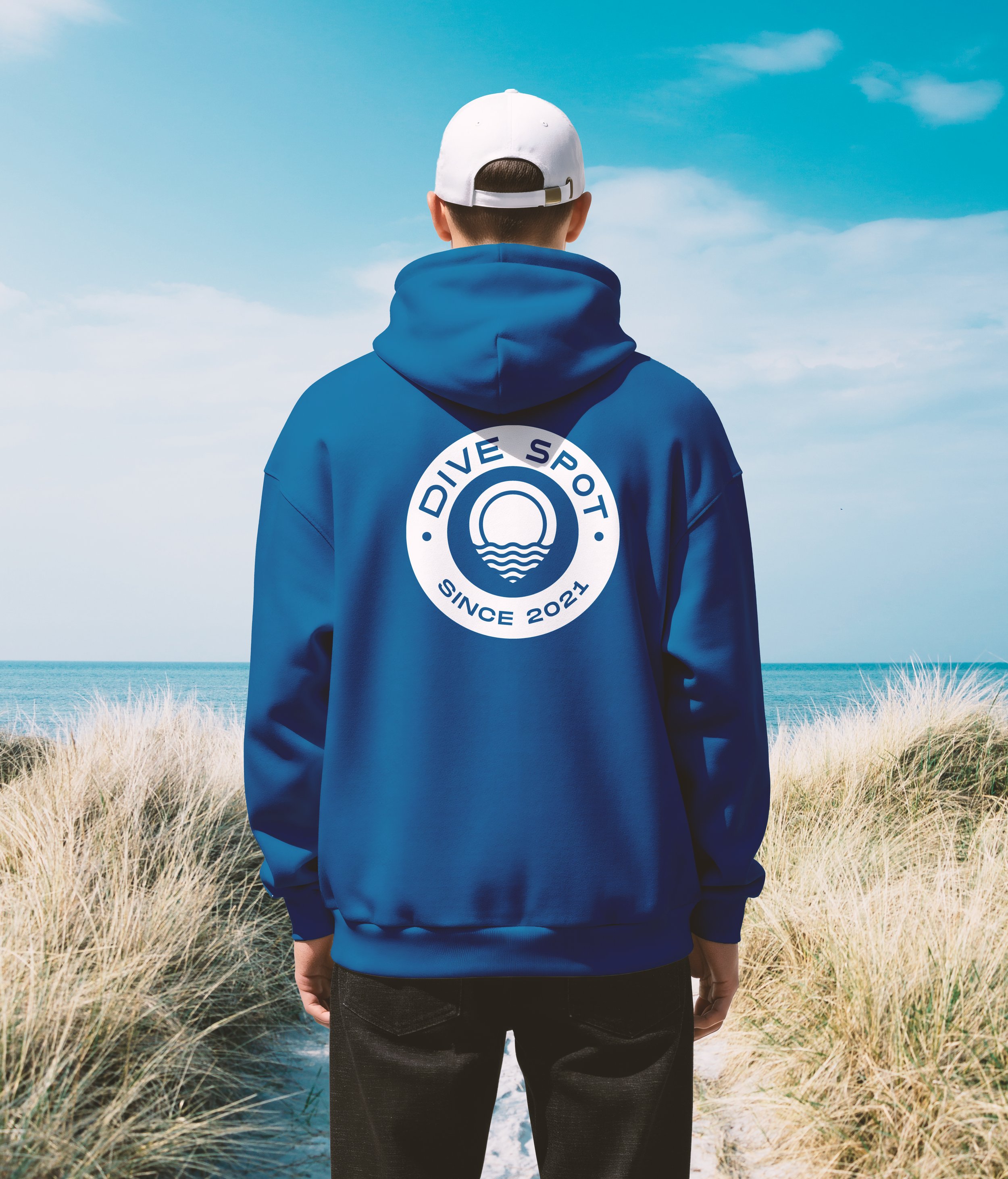 Persona de espaldas observando el mar y la playa, lleva una sudadera azul con un logotipo de deportes acuáticos, gorra blanca y pantalones oscuros, rodeado de pasto seco y cielo azul con nubes.