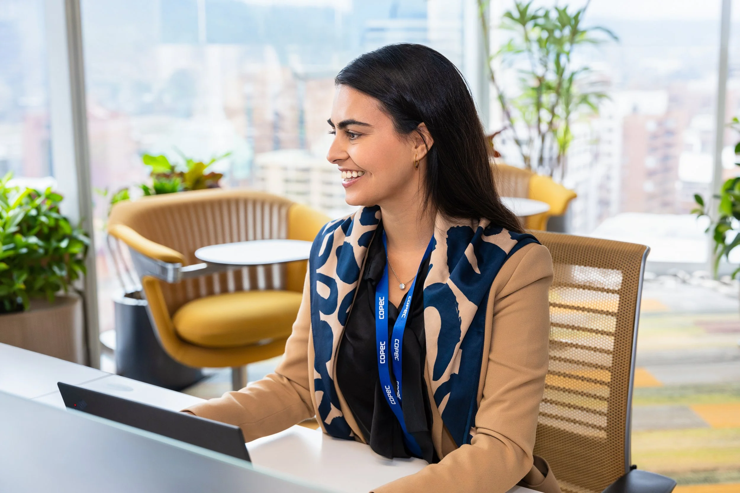 Mujer sonriendo en una oficina moderna con plantas y ventanas grandes al fondo, sentada frente a una computadora portátil.