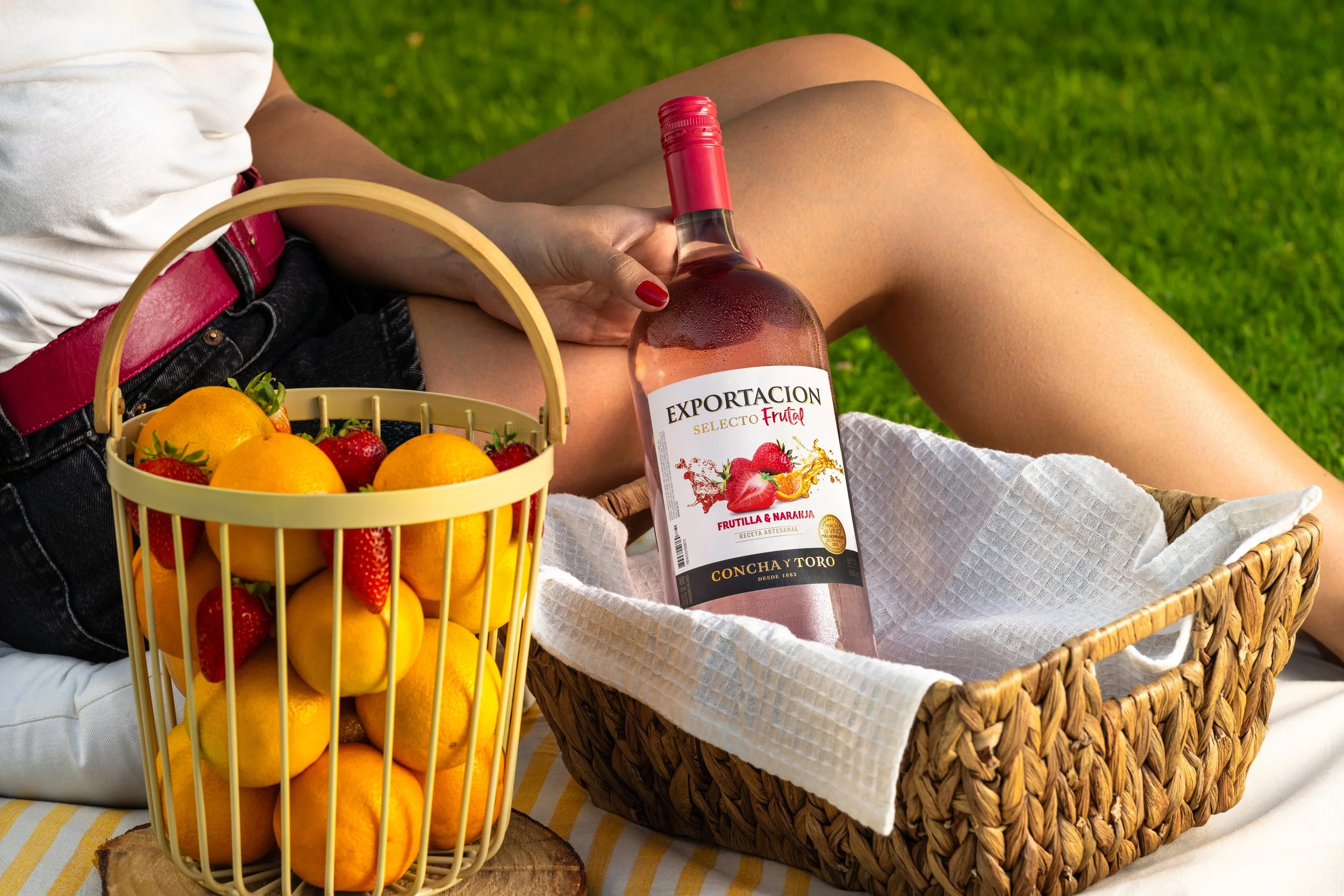 Persona sosteniendo una botella de vino rosado con una canasta llena de frutas como naranjas y fresas, en un picnic sobre césped.