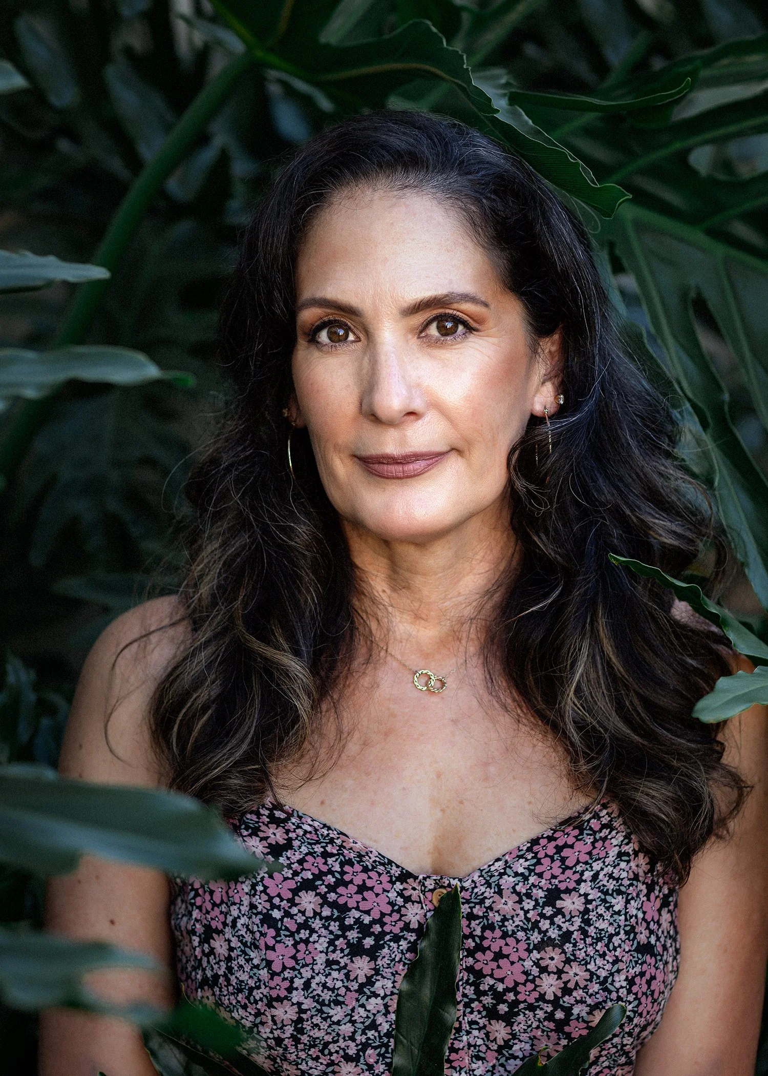 Life coach branding portrait of a woman with long, wavy dark hair, smiling softly among large green tropical leaves, photographed for her coaching website by Vera Gosic Photography in Long Beach, California.