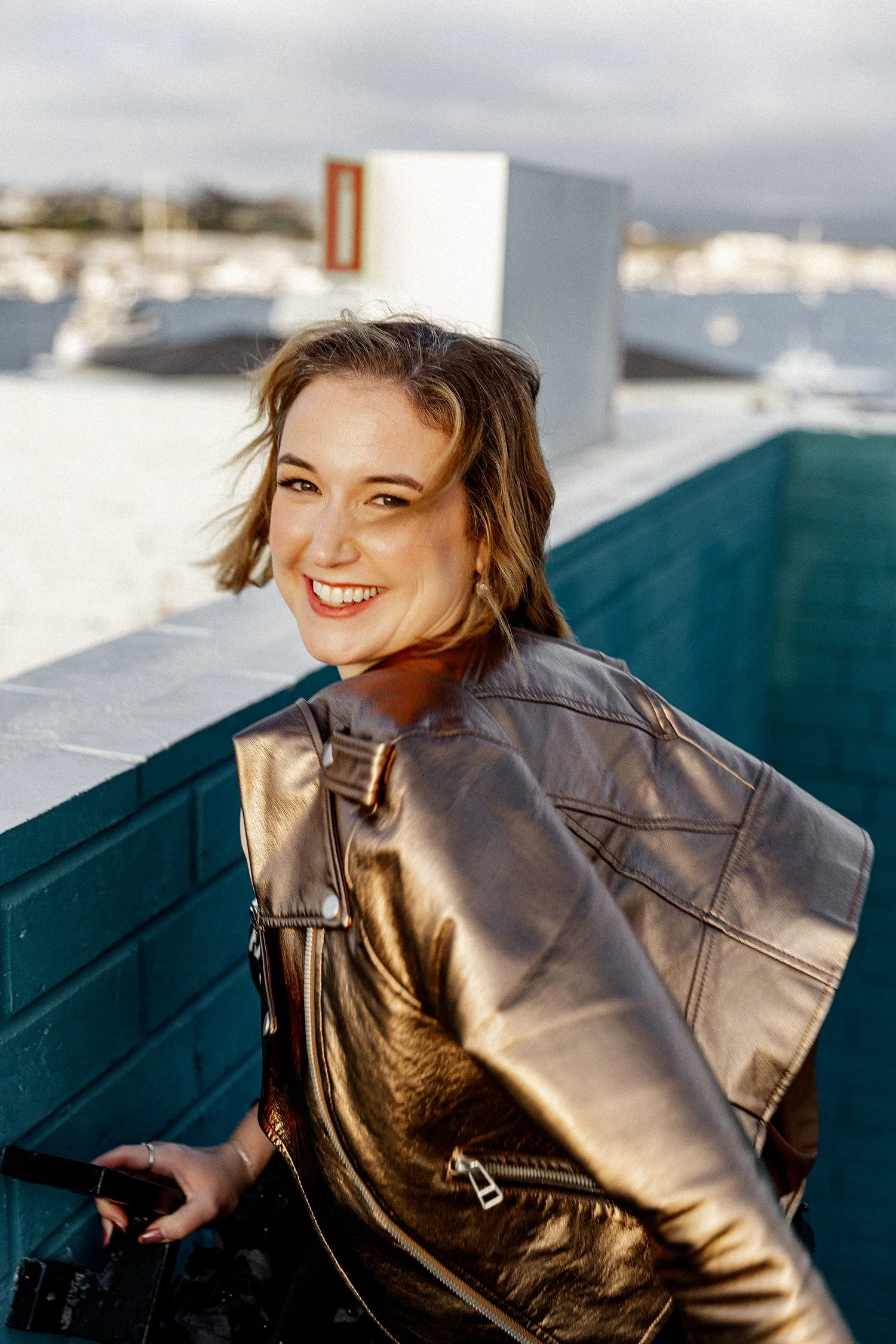 Personal stylist branding photo of a smiling woman crossing a palm-lined street at sunset in Newport Beach, wearing a bronze metallic leather jacket, a sheer black floral tulle skirt, black heeled boots, and carrying a black handbag with rattan trim.