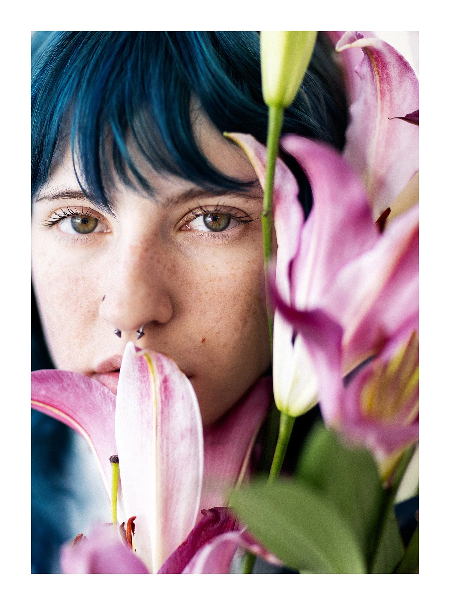 Close-up fine art portrait of a person with dark blue hair, hazel eyes, freckles, and a septum piercing, partly hidden behind pink stargazer lilies.