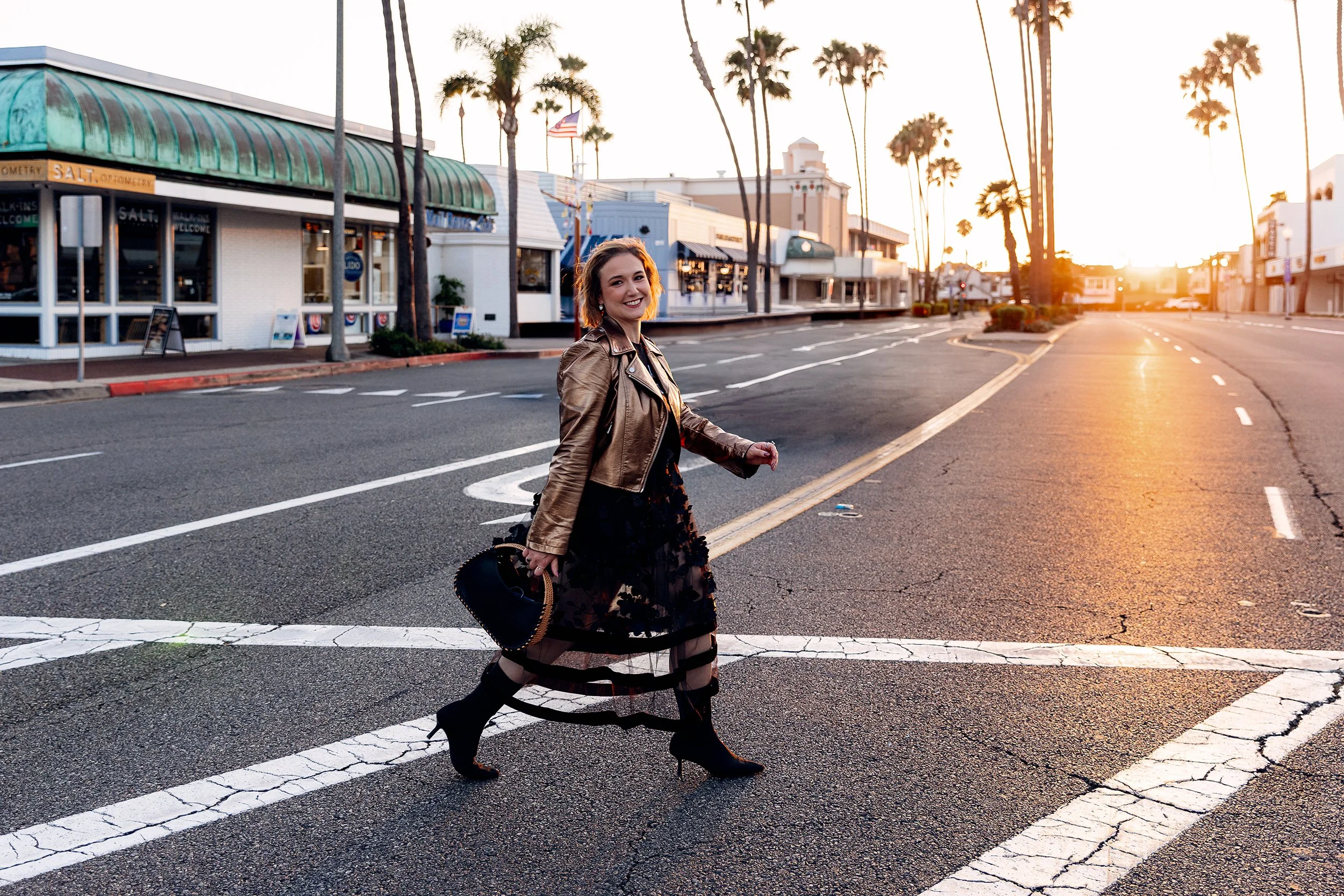 Personal stylist branding photo of a smiling woman crossing a palm-lined street at sunset in Newport Beach, wearing a bronze metallic leather jacket, a sheer black floral tulle skirt, black heeled boots, and carrying a black handbag with rattan trim.