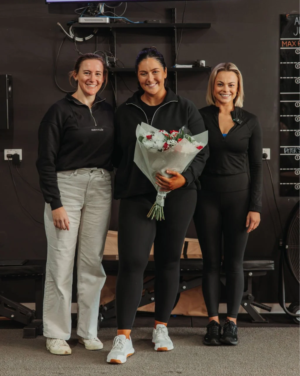 Three Personal Trainers together indoors, smiling, with the woman in the center holding a bouquet of flowers.
