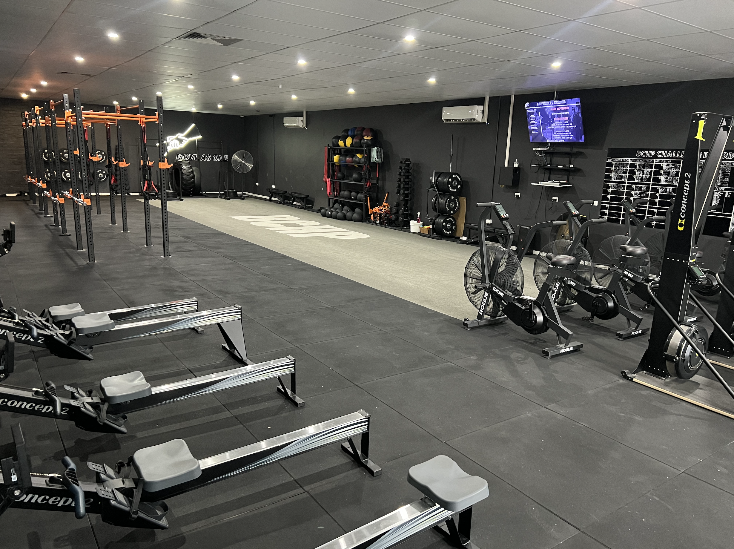 Empty gym with workout equipment including rowers, assault bikes, Ski Ergs, Cable Machines. Squat Racks, weight racks, medicine balls, and a large TV screen on the wall.