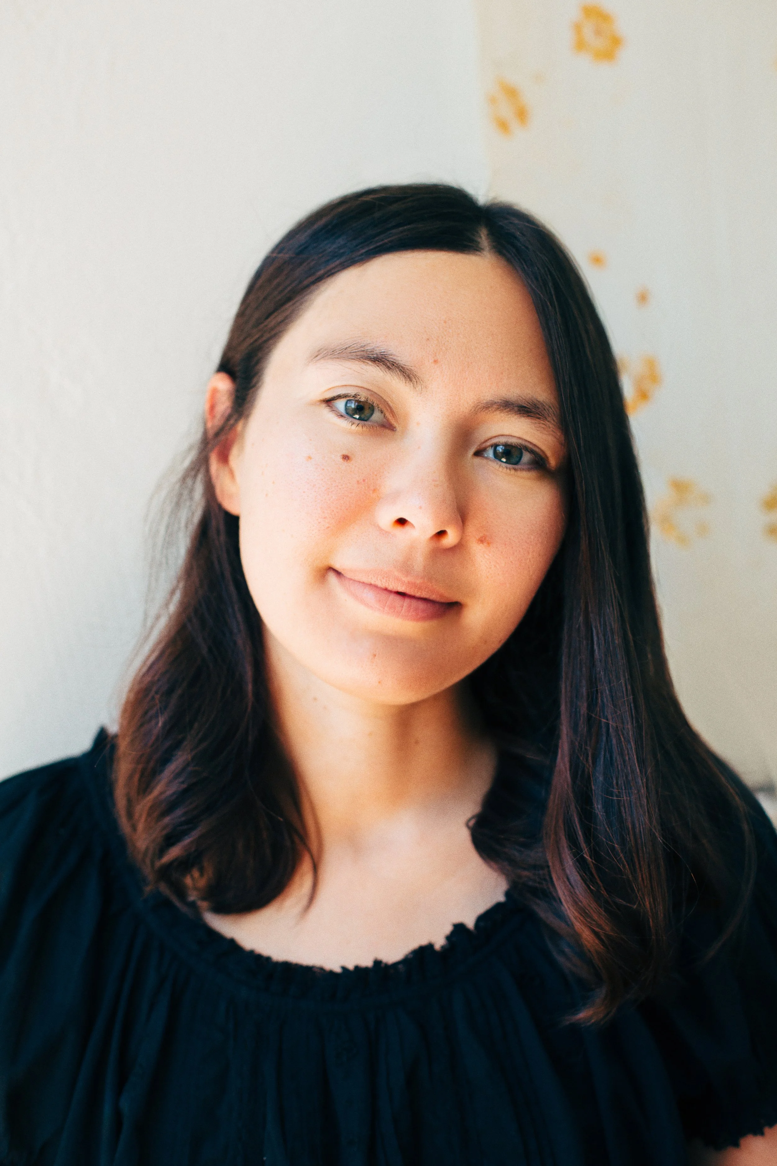 Close-up portrait of a young woman with brown hair, blue eyes, and a subtle smile, wearing a black top, standing against a light-colored wall with yellow paint splatters.