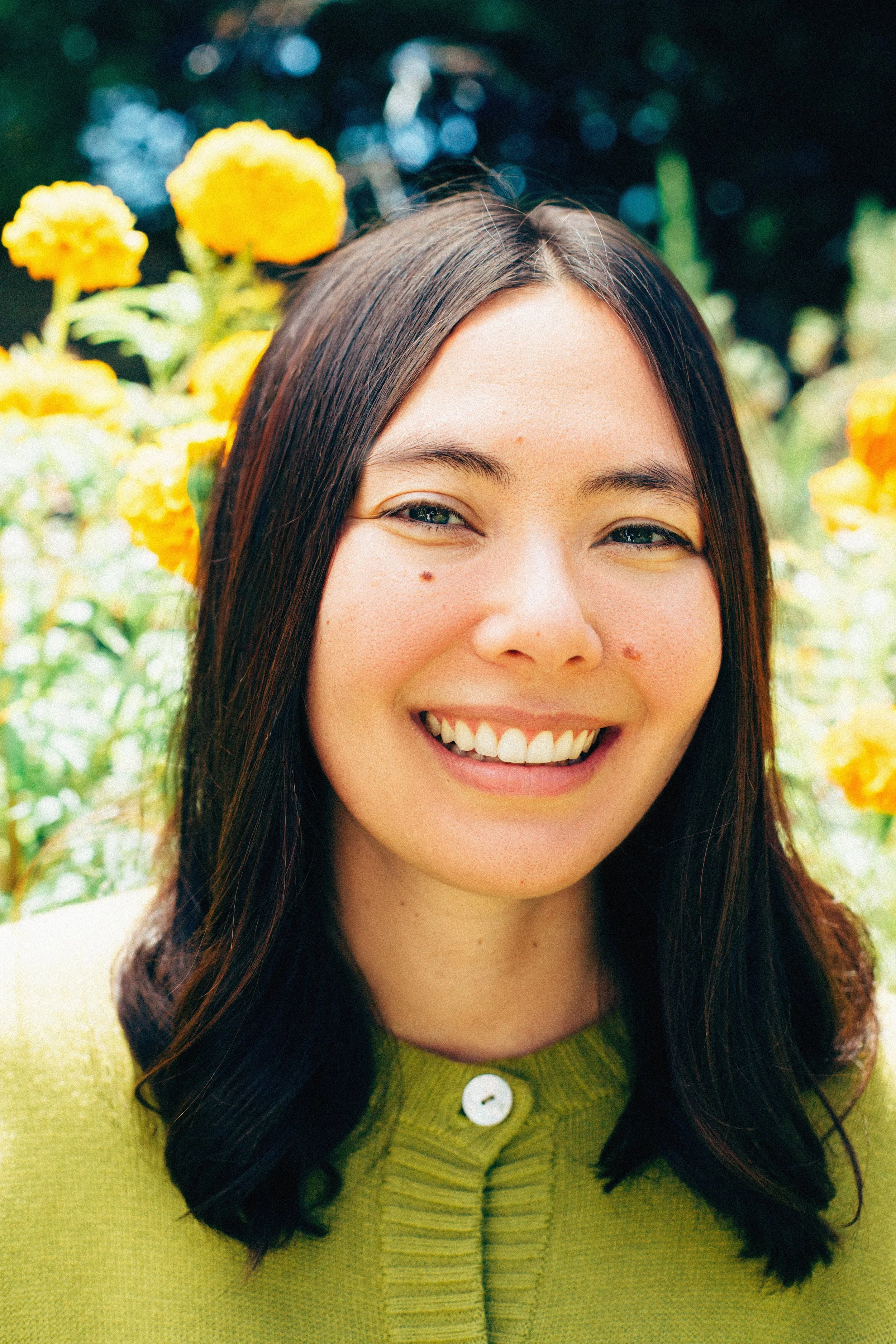 Close-up of a young woman with long dark hair smiling in a garden with yellow flowers in the background.