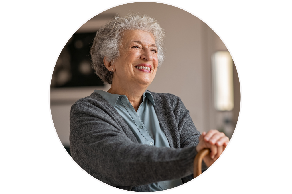 An elderly woman with curly grey hair smiling, wearing a blue shirt and a grey cardigan, holding a wooden cane, sitting indoors in a well-lit room. She is content and satisfied receiving quality aged care services, from the comfort of her home.