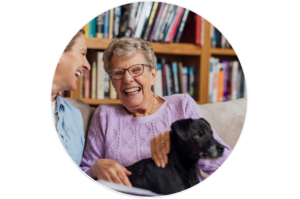 Two women, one elderly and one younger, laughing together on a couch with a small black dog, in a room with a bookshelf in the background. The elderly woman. is happy and relaxed knowing she receives consistent and trustworthy care at home.