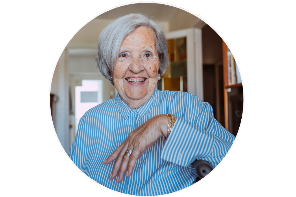 An elderly woman with short gray hair smiling, wearing a blue and white striped shirt, seated indoors with a bright background. She's relaxed at home receiving premium aged care services, in South Melbourne.