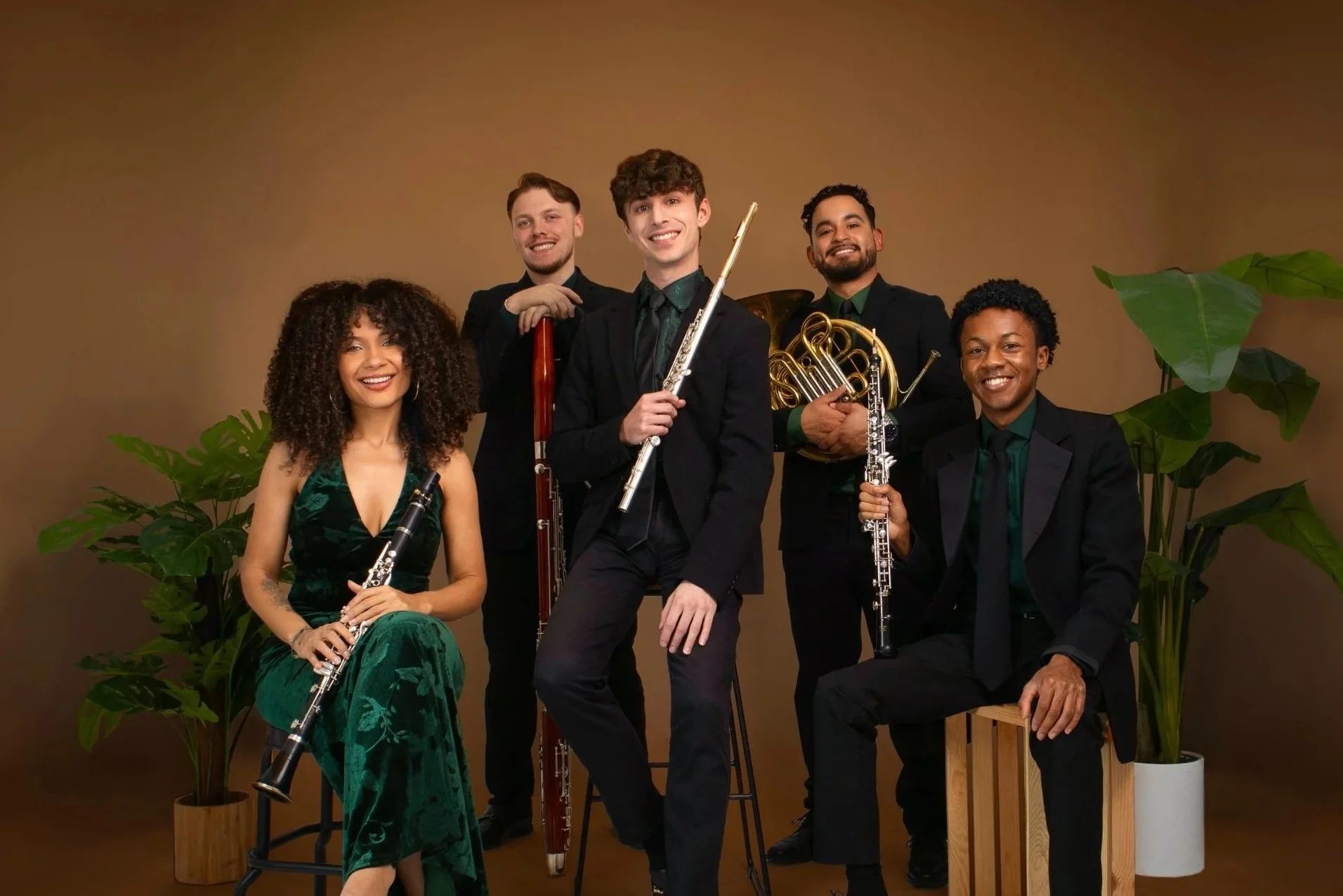 Group of six musicians dressed in formal attire posing with their musical instruments, surrounded by green plants in a studio with a brown background.