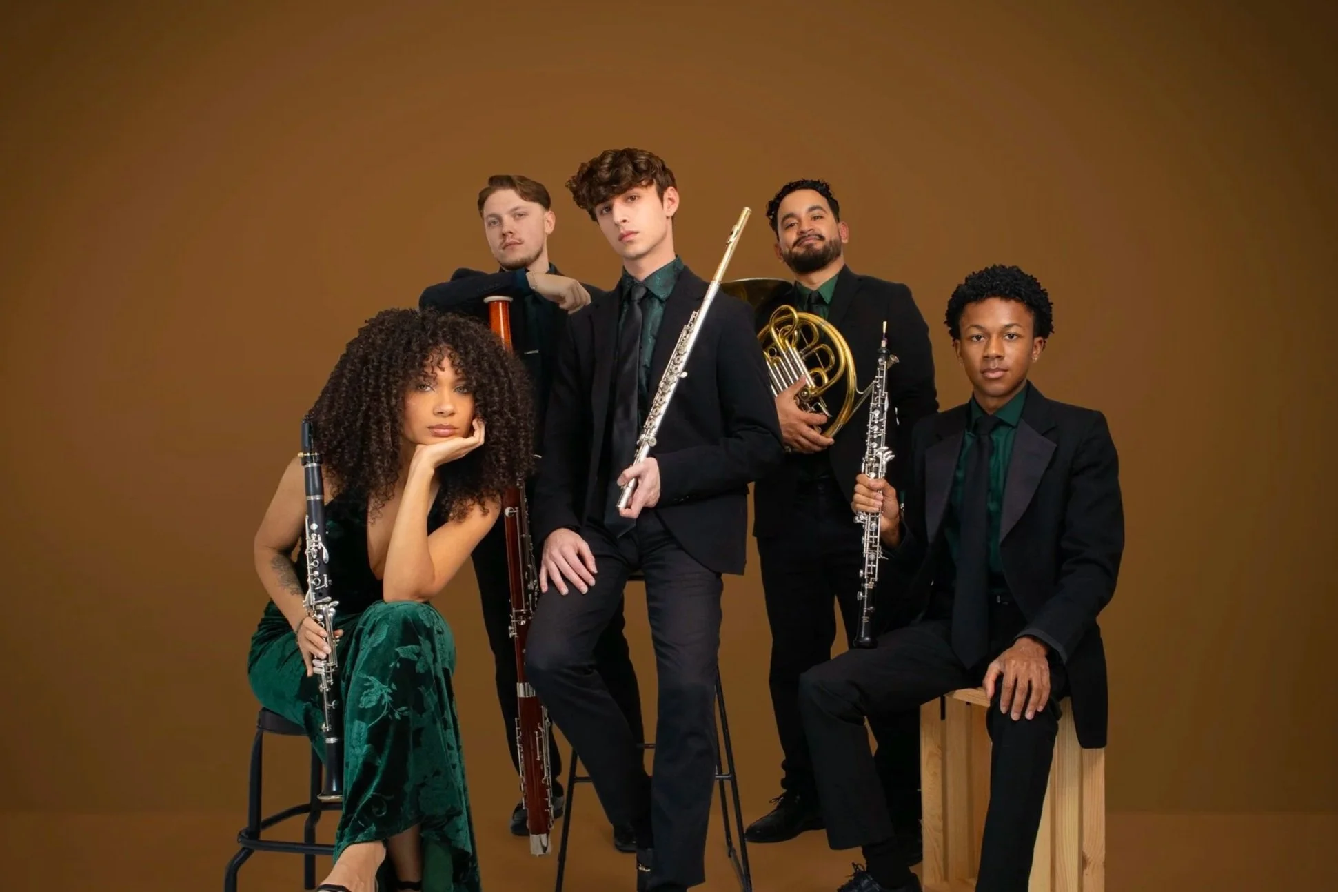 A group of five musicians holding various musical instruments against a plain brown background. One woman sits on a stool holding a flute, all wearing formal black attire with green accents.