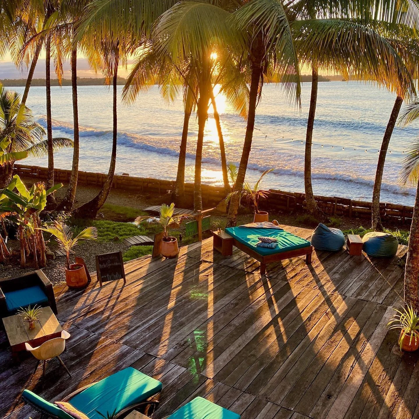 Sunset view overlooking a beach from a wooden deck with lounge chairs, bean bags, potted plants, and palm trees.