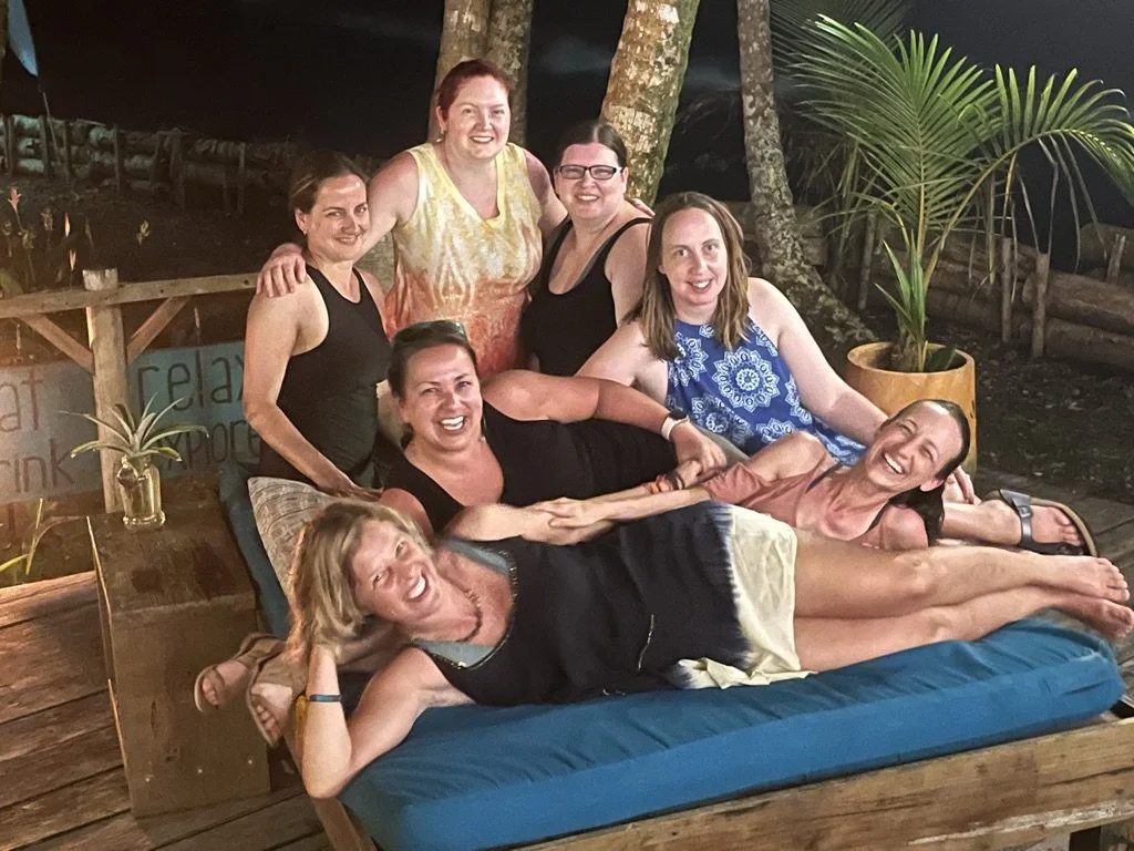 Group of seven women at night outdoors, some lying on a lounging bed, smiling and enjoying each other's company, surrounded by trees and potted plants.