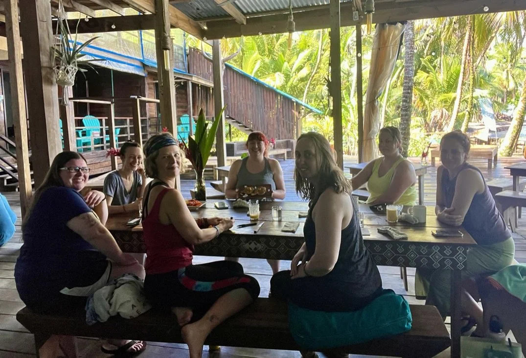 A group of seven women sitting around a wooden table at an outdoor tropical restaurant, smiling and enjoying drinks and food. The setting has lush green foliage in the background and a wooden structure with blue chairs.