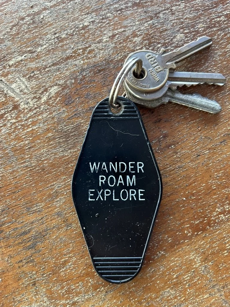 A set of keys with a black keychain that reads 'WANDER ROAM EXPLORE' on a wooden surface.