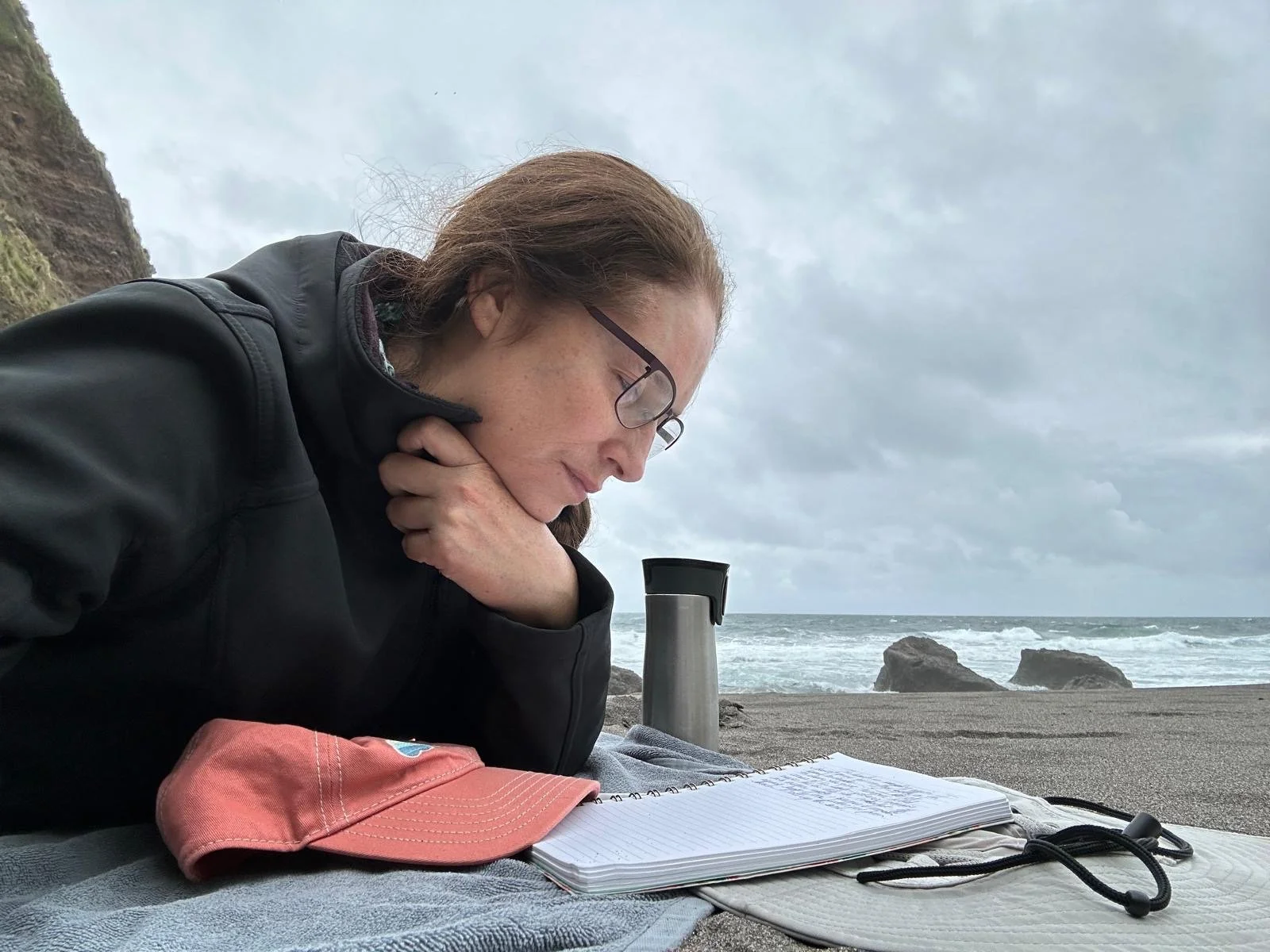 Woman with glasses reading a notebook on a beach under cloudy sky, with rocks in the ocean in the background.