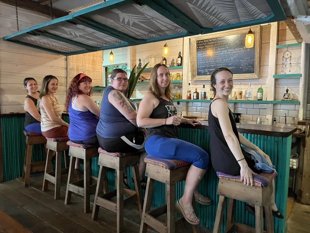 Six women sitting on bar stools at a rustic bar with a wooden interior, smiling and enjoying drinks.