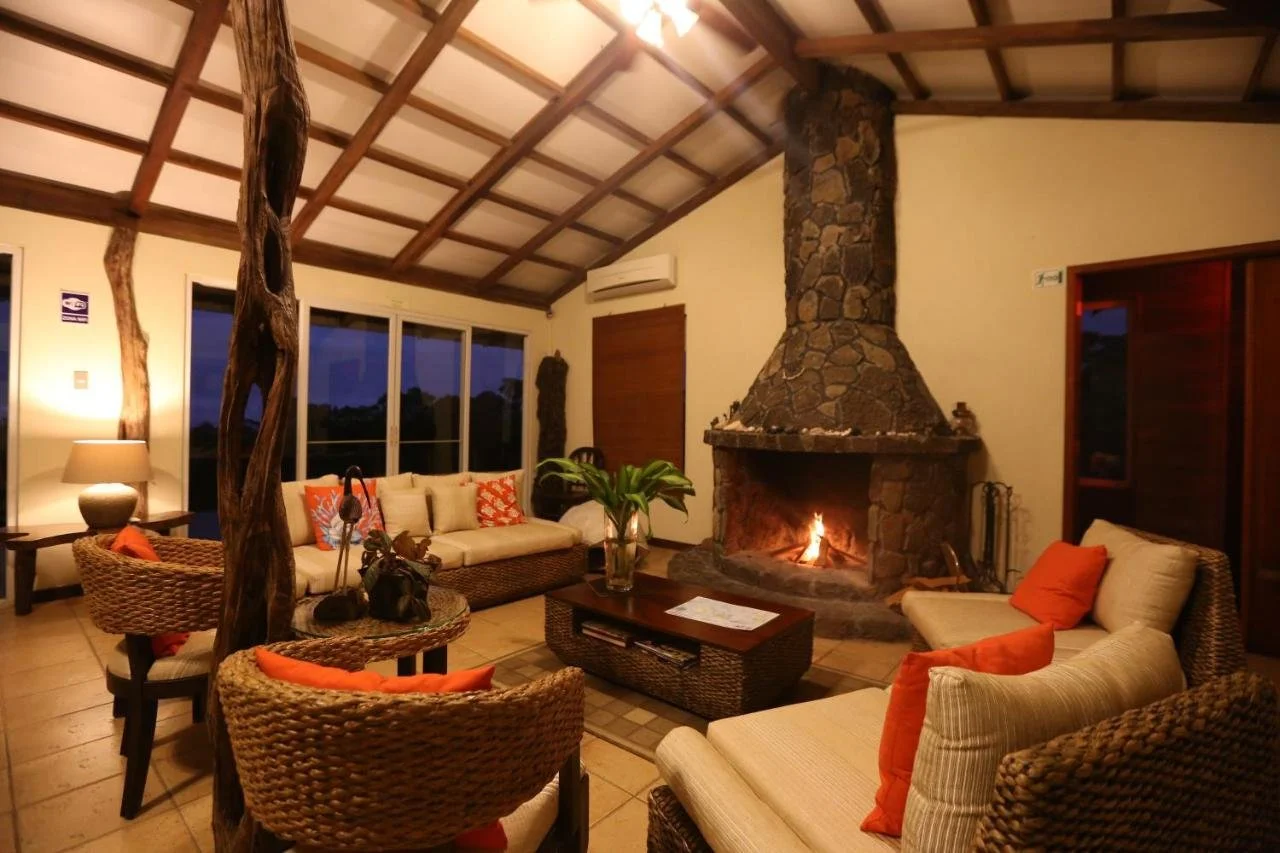 Cozy living room with rattan furniture, orange cushions, and a large stone fireplace with a fire burning. There is a wooden coffee table with a plant in a glass vase, a side table with a lamp, and large windows showing a dark sky outside. The ceiling