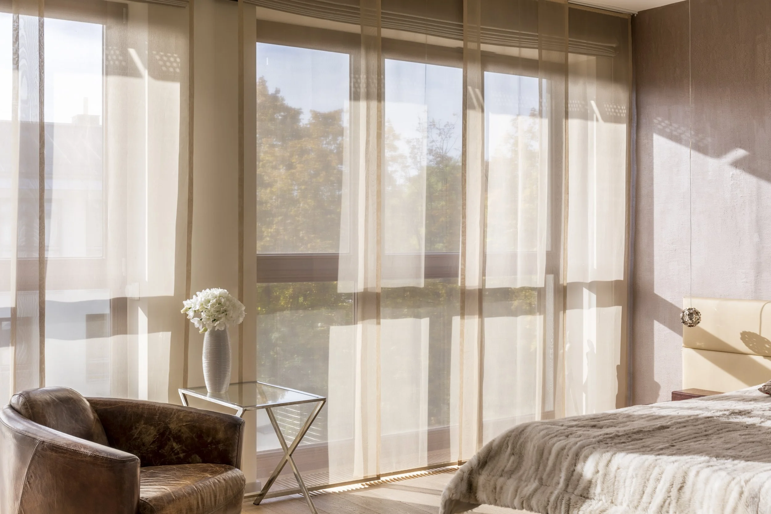 Bright bedroom interior with sheer curtains, a bed with a beige headboard and furry blanket, a glass side table with a white vase of white flowers, and a brown leather armchair.