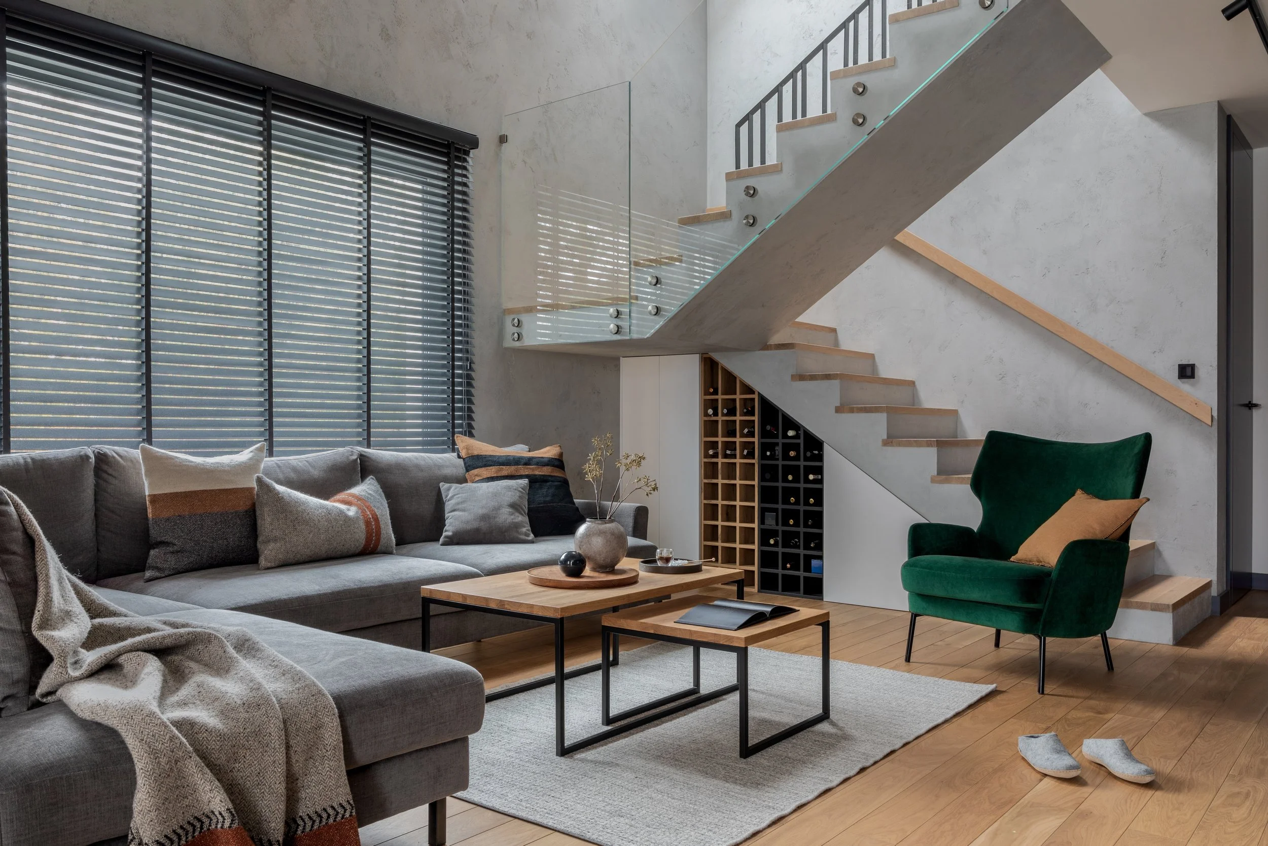 Modern living room with gray sectional sofa, colorful pillows, wooden coffee tables, green armchair, and staircase with glass railing and built-in wine storage.