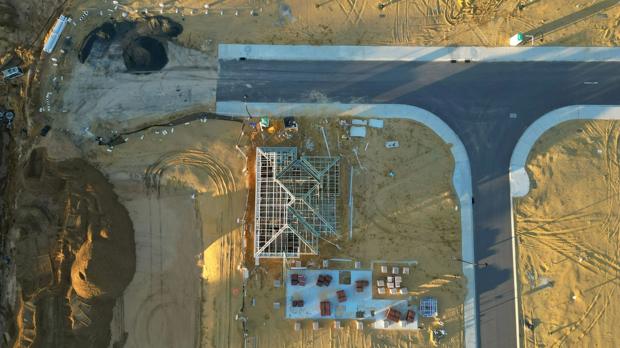 Aerial view of a construction site showing a partially built house with a metal framework, a newly paved road with a curve, and surrounding dirt and construction materials.
