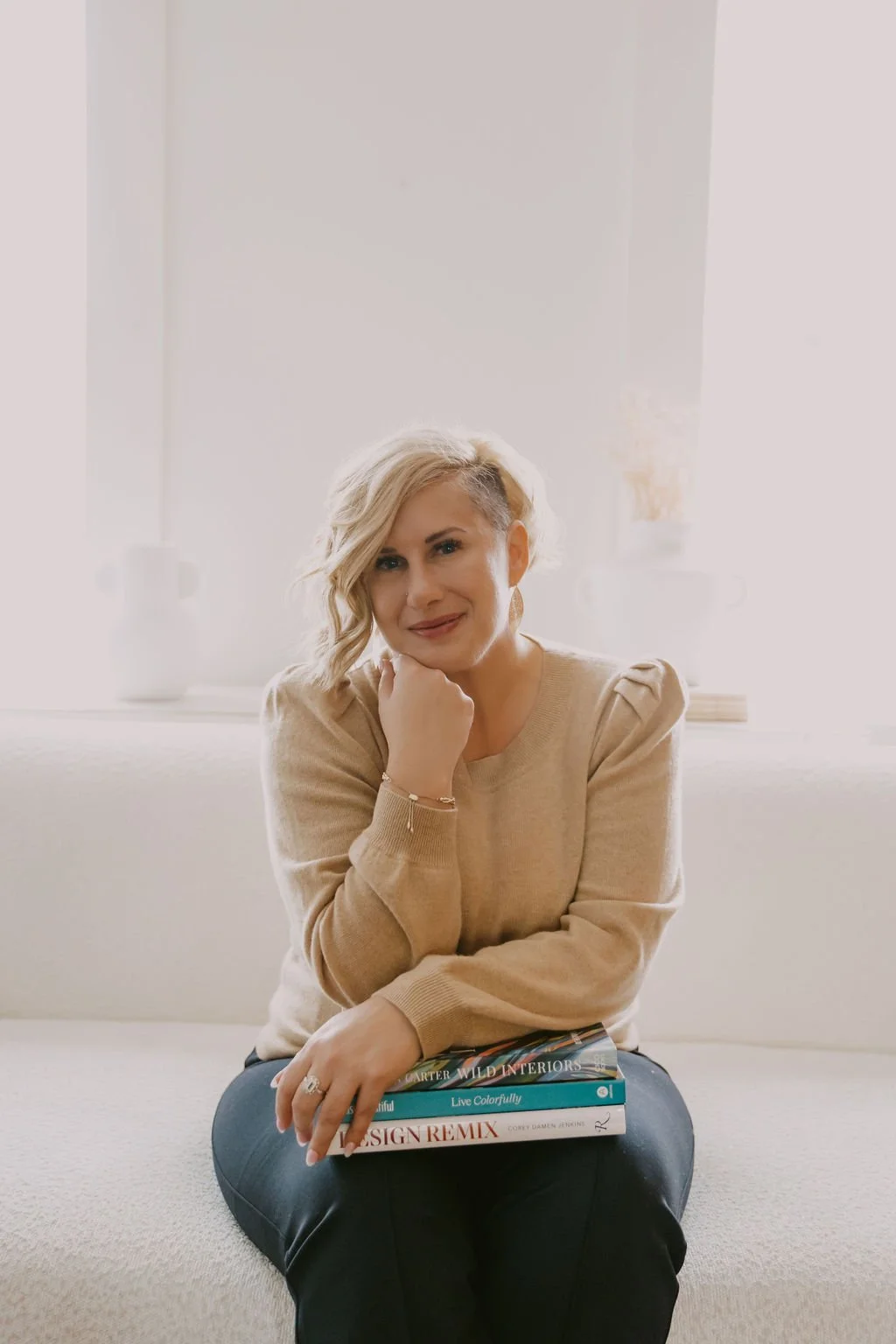 A woman with short blonde hair and a warm smile sitting on a light-colored sofa, resting her chin on her hand, holding a stack of books, in a bright room with white walls and decorative vases in the background.