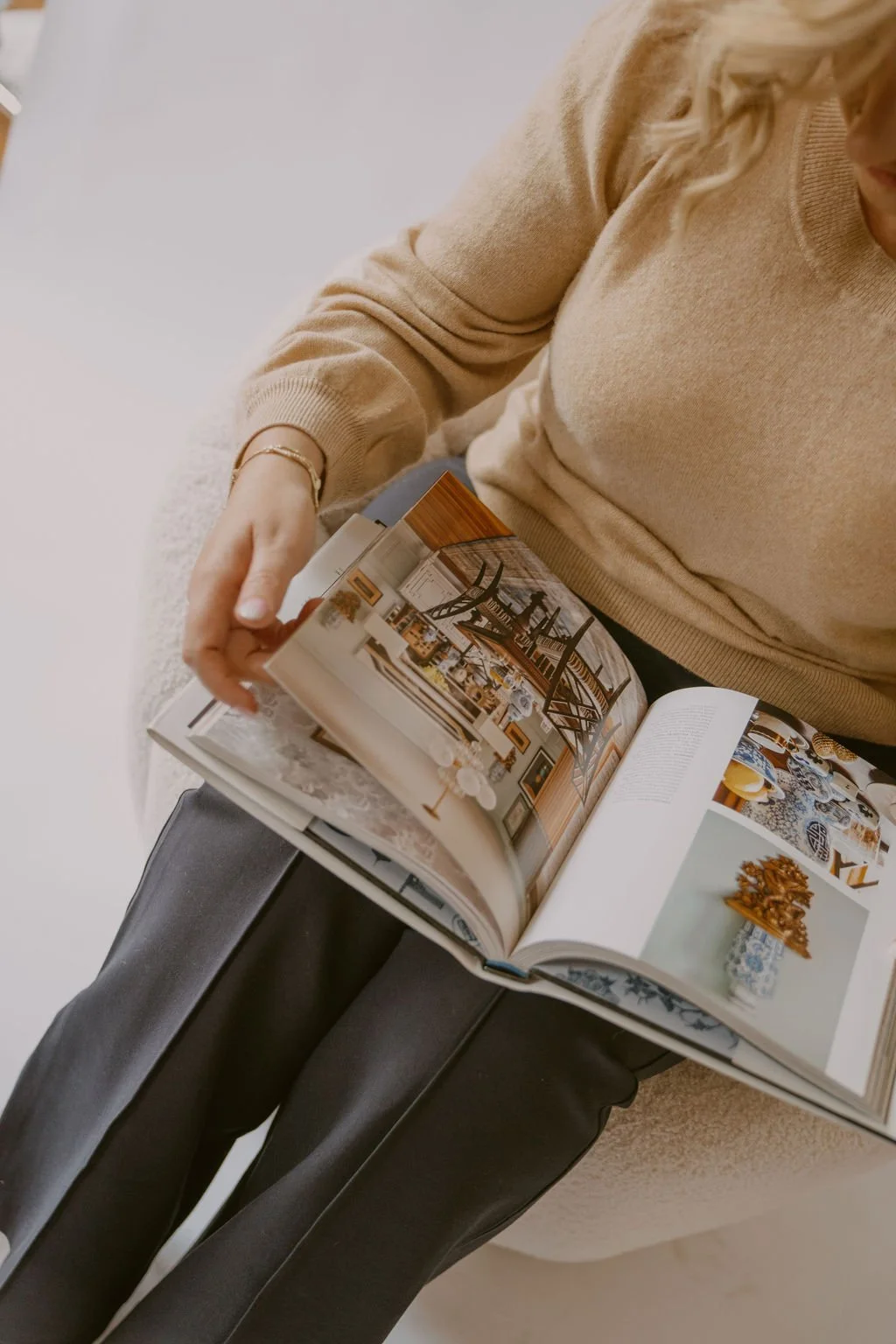 A woman in a beige sweater and dark pants sitting on a chair, reading a magazine that features interior design and home decor images.