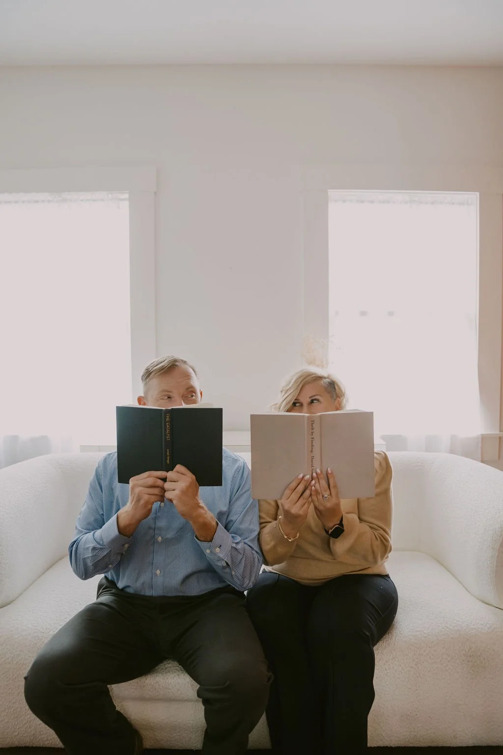 A man and a woman sitting on a white couch reading books, with bright light coming through two windows behind them.