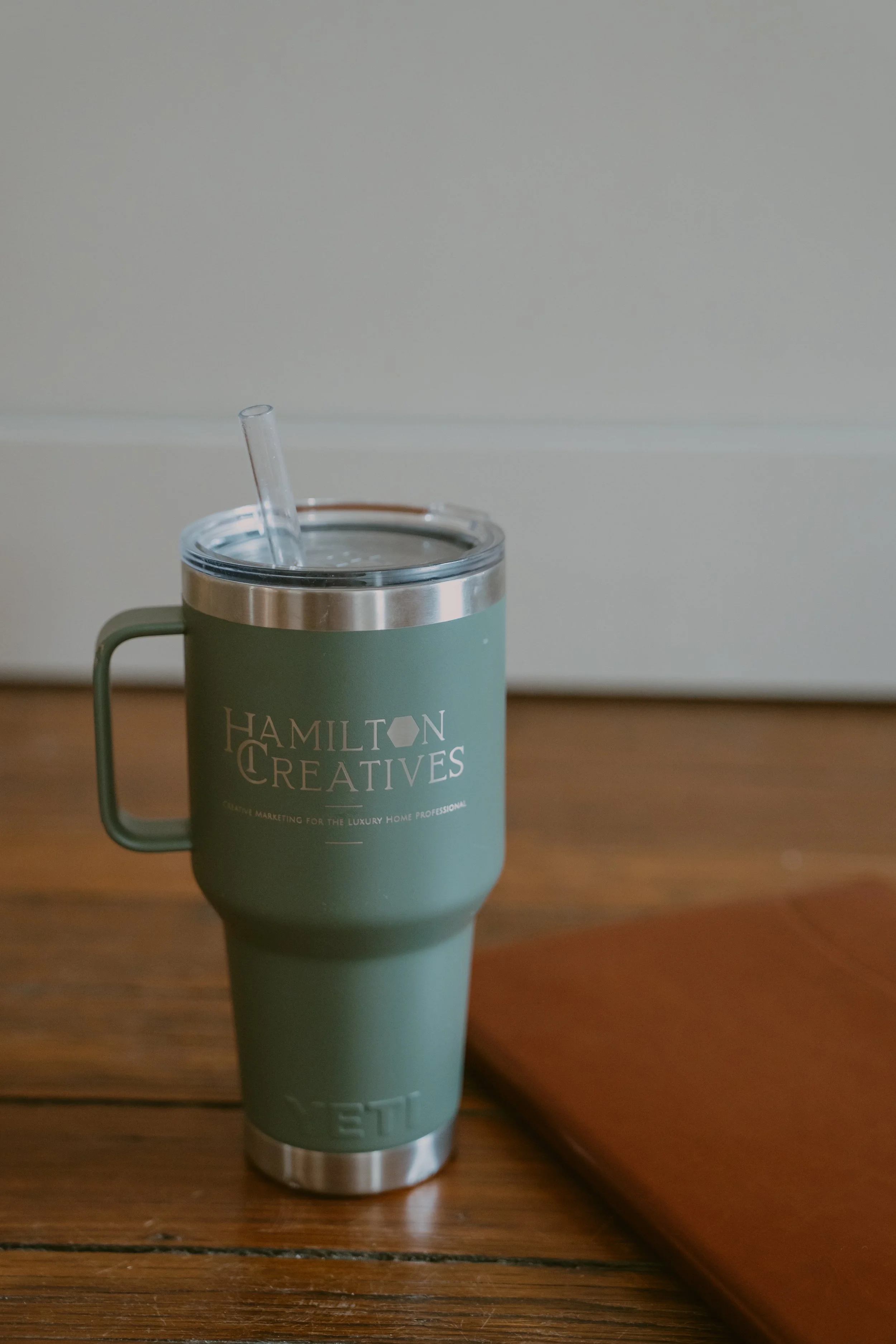 A green travel tumbler with a transparent straw on a wooden table. The tumbler has the text 'Hamilton Creatives' printed on it.