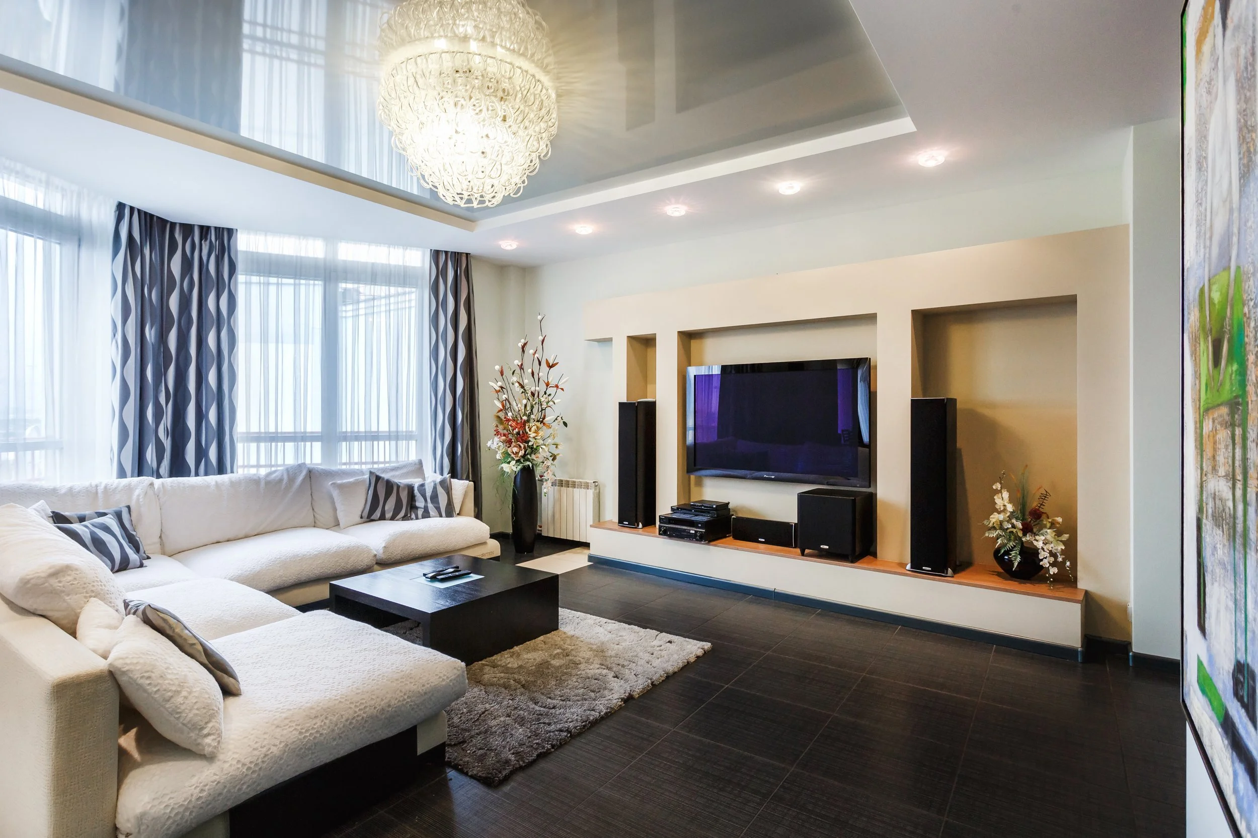 Living room with white sectional sofa, dark wood coffee table, patterned curtains, large window, flat-screen TV, surround sound speakers, floral arrangements, chandelier, and a multicolored abstract painting on the wall.