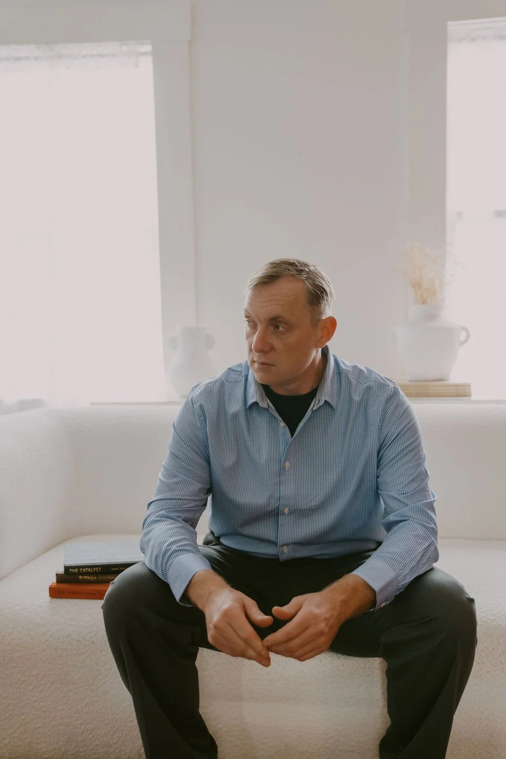 A middle-aged man in a blue checked shirt sitting on a white sofa, with a stack of books beside him, in a bright room with large windows.