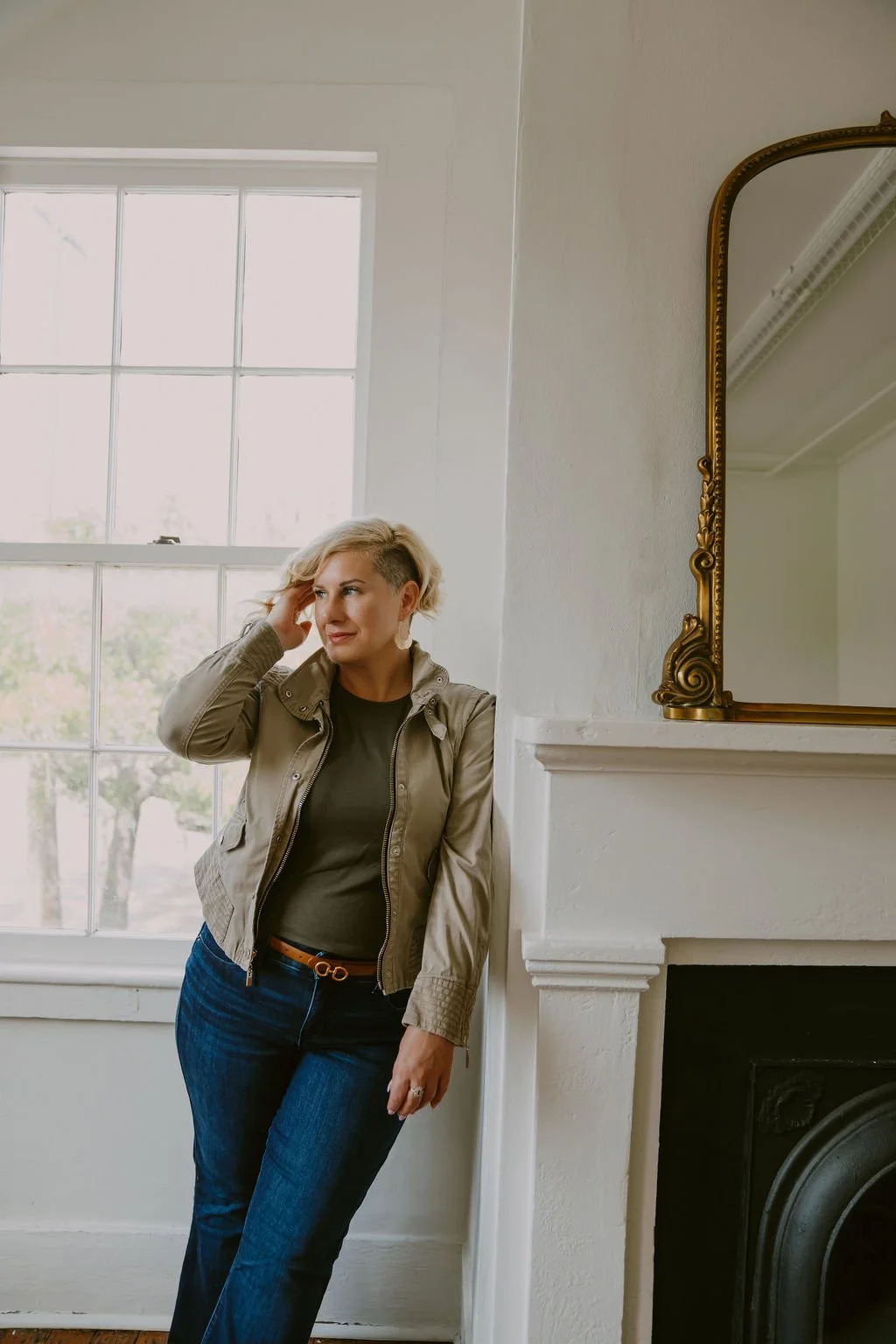 A woman with short blonde hair, wearing a beige jacket, black shirt, and blue jeans, stands next to a white fireplace with a gold ornate mirror on the mantel. She is indoors near a large window, looking thoughtful while touching her head.
