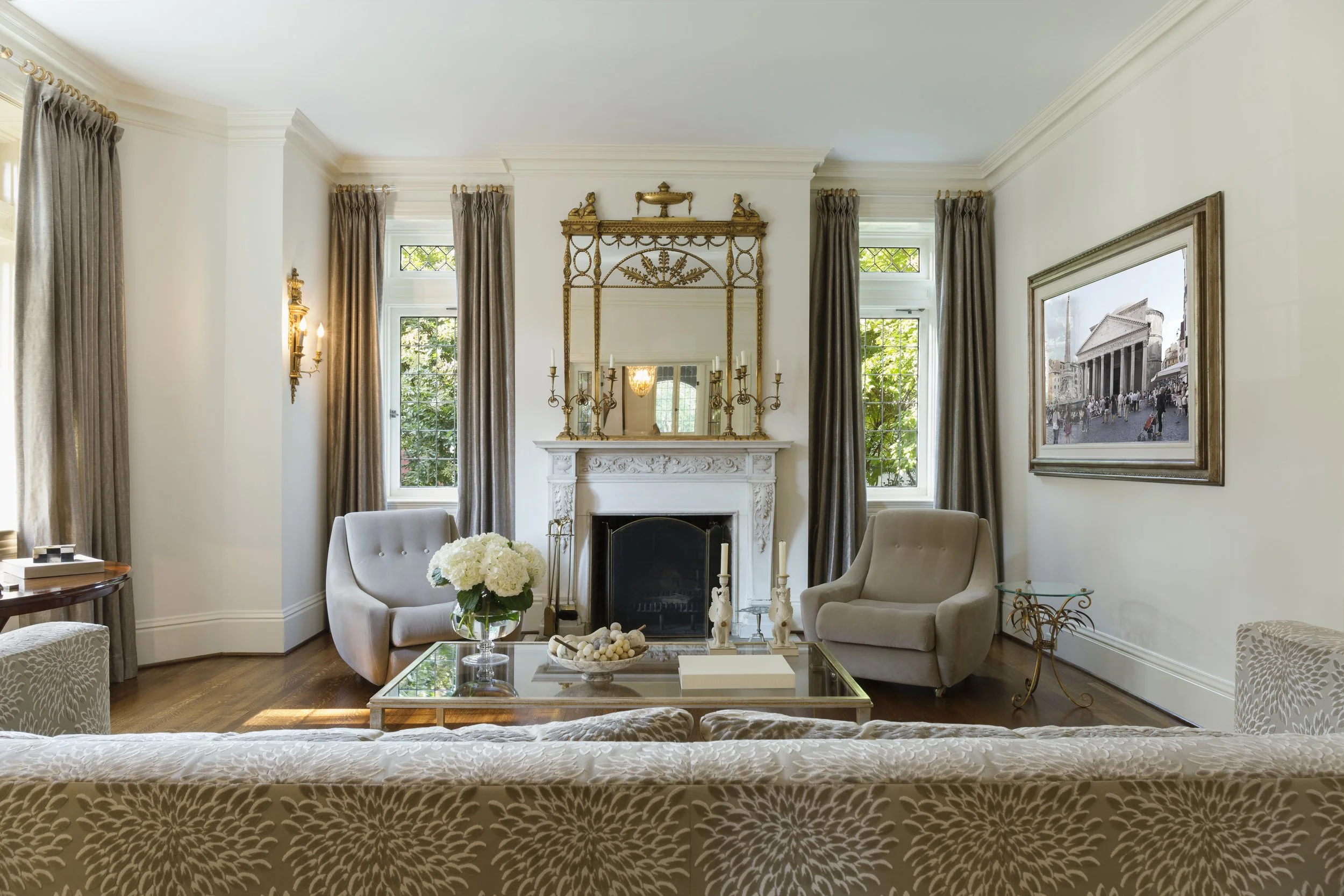 Elegant living room with white walls, a white ornate fireplace, two beige armchairs near the fireplace, a glass coffee table with white flowers and decor, dark curtains framing three windows with greenery outside, a large framed painting of a historic building, and ornate gold accents on the mirror, candle holders, and side table.